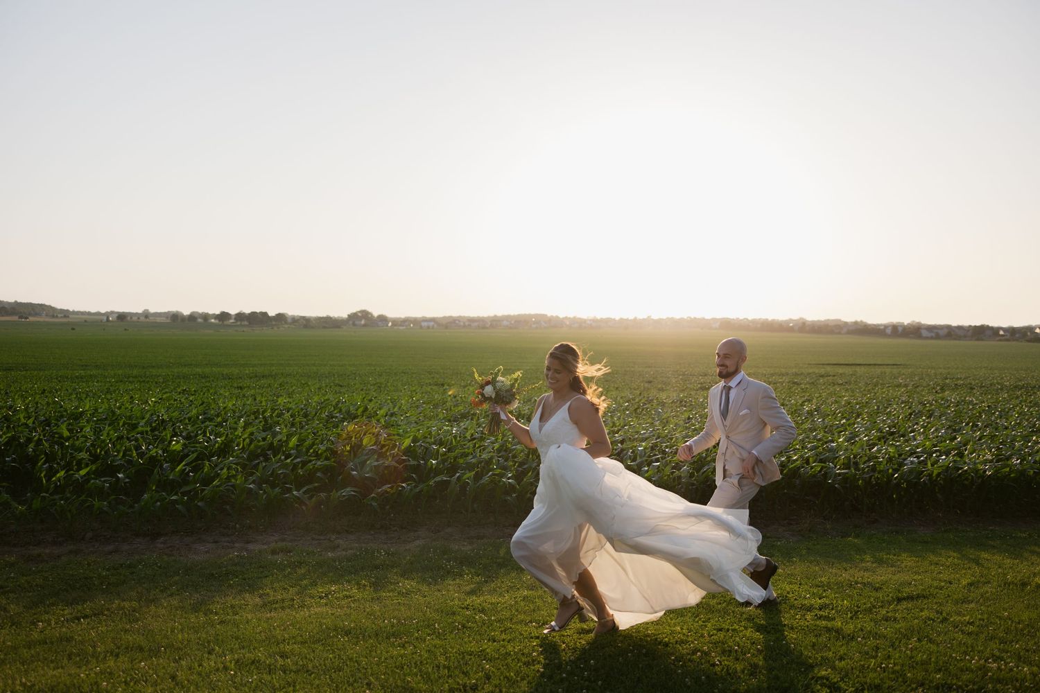 A couple dances together in a flowing white dress at sunset in a green rural field with sunflowers nearby.