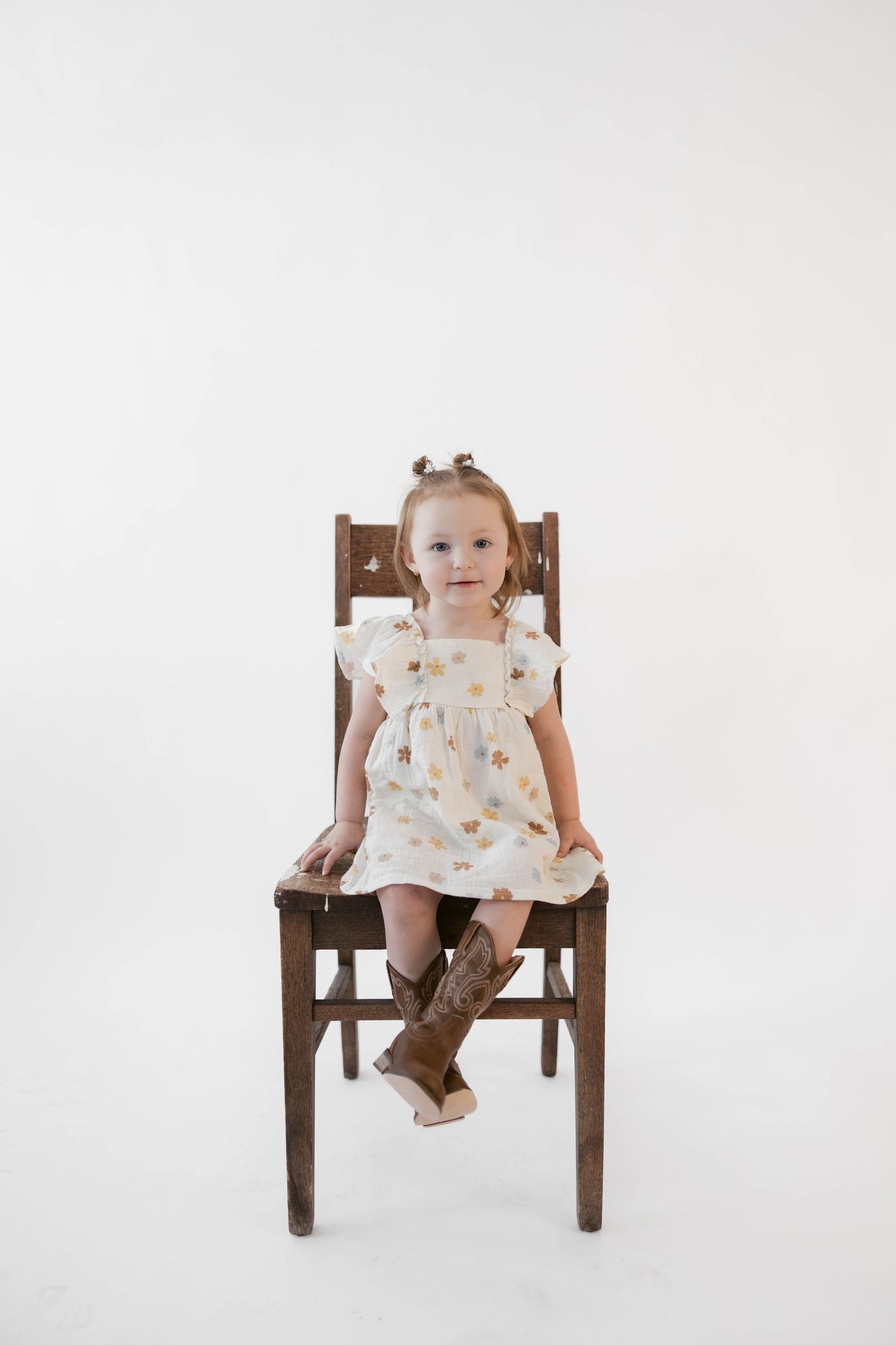 Little girl in floral dress and boots sitting on wooden chair against white background.