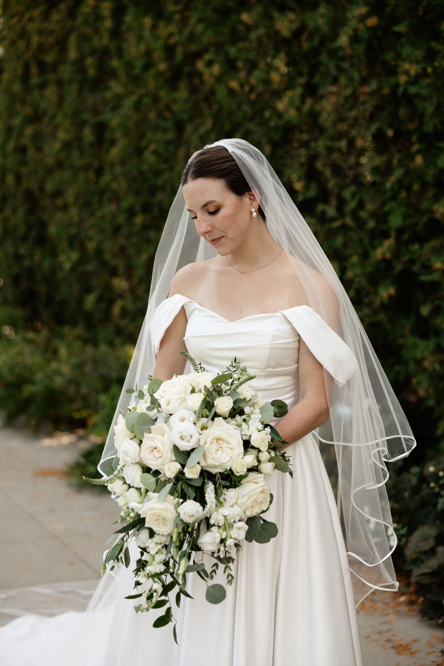 A bride in a strapless white wedding dress holds an elegant bouquet of white roses and eucalyptus against a garden backdrop.