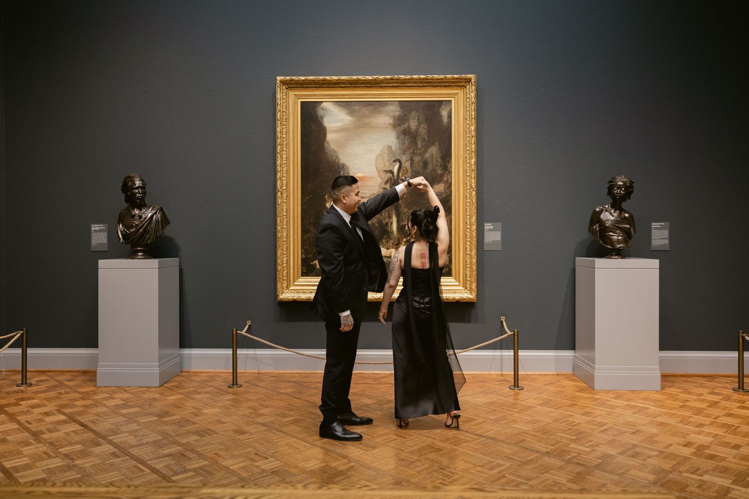 A couple poses romantically in an art gallery with gold-framed paintings on dark walls.
