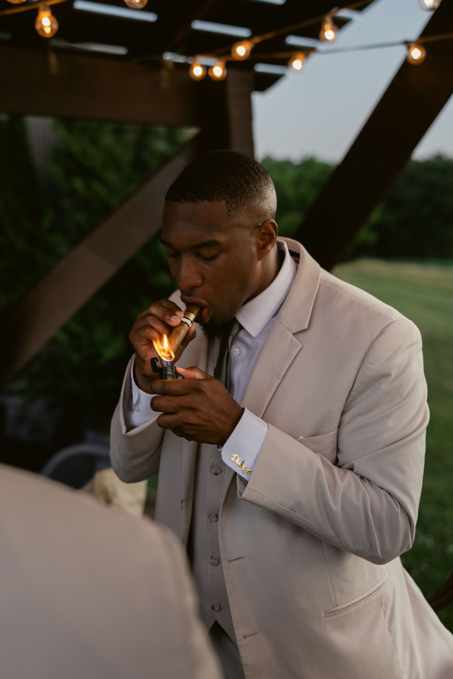 Person in light gray suit lighting a cigar at an outdoor venue with string lights overhead.