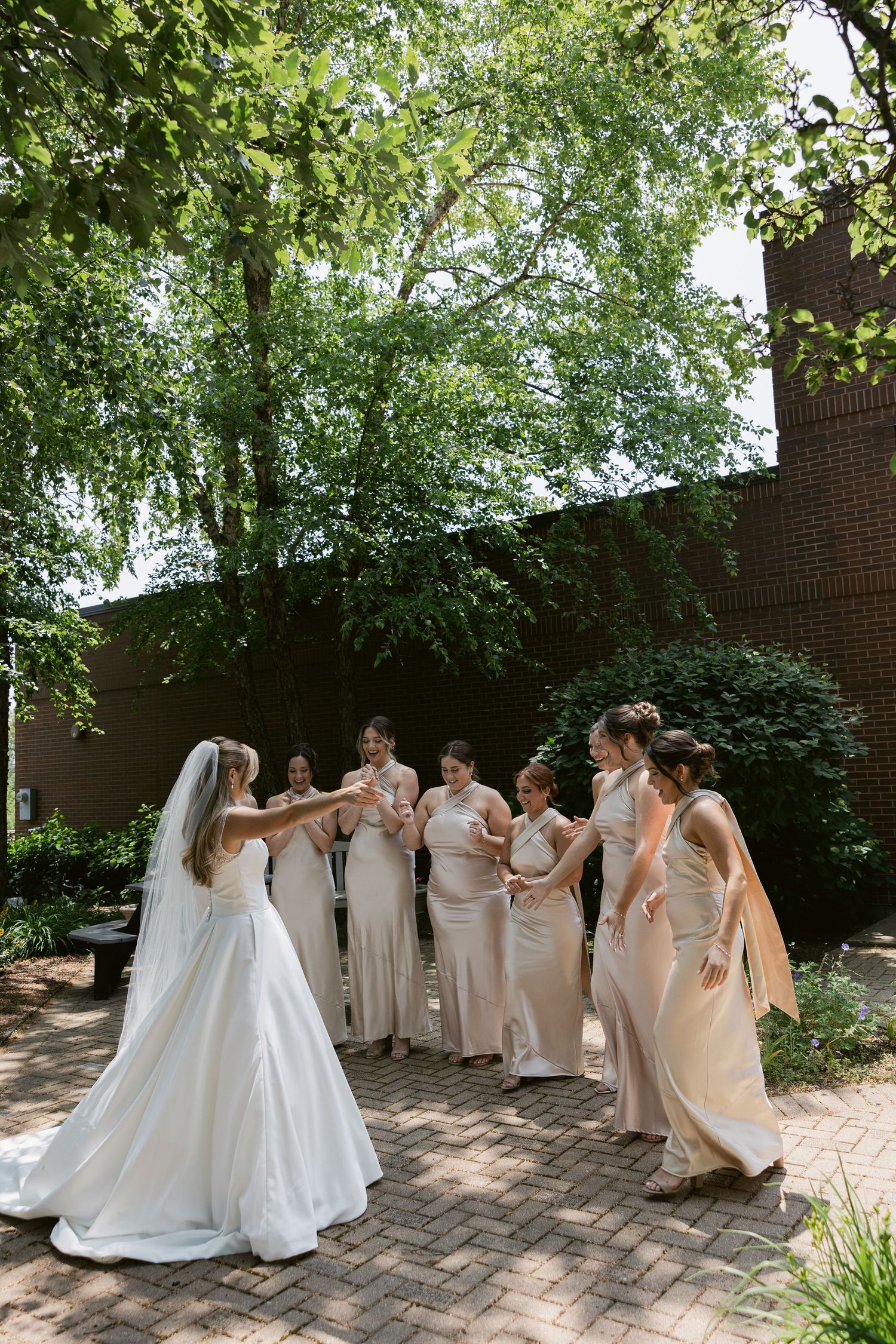 Bridal party in flowing champagne-colored dresses gathers together on a brick patio surrounded by lush greenery.