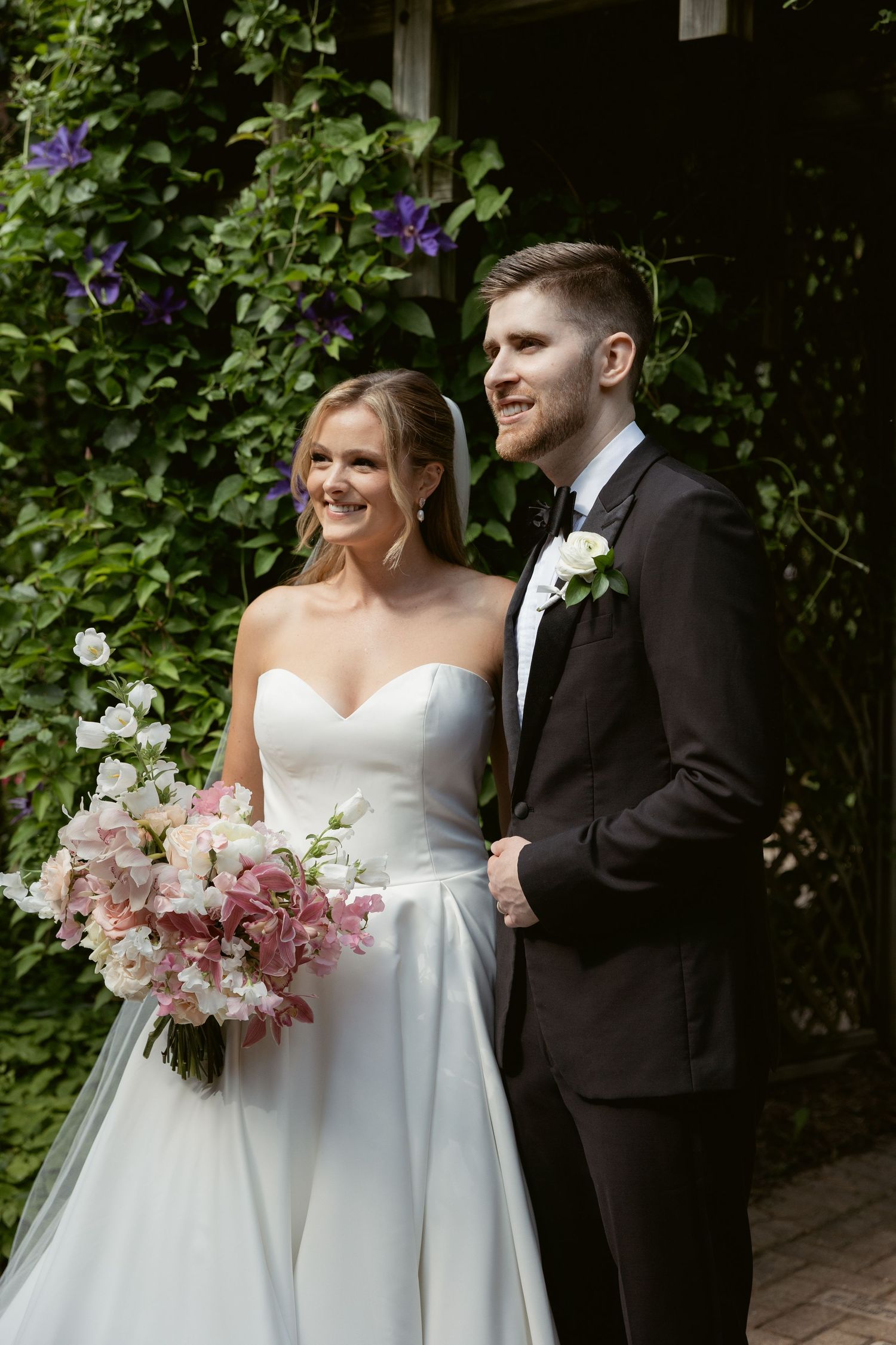 Wedding couple posing in garden with purple flowers, bride holding pink and white bouquet.