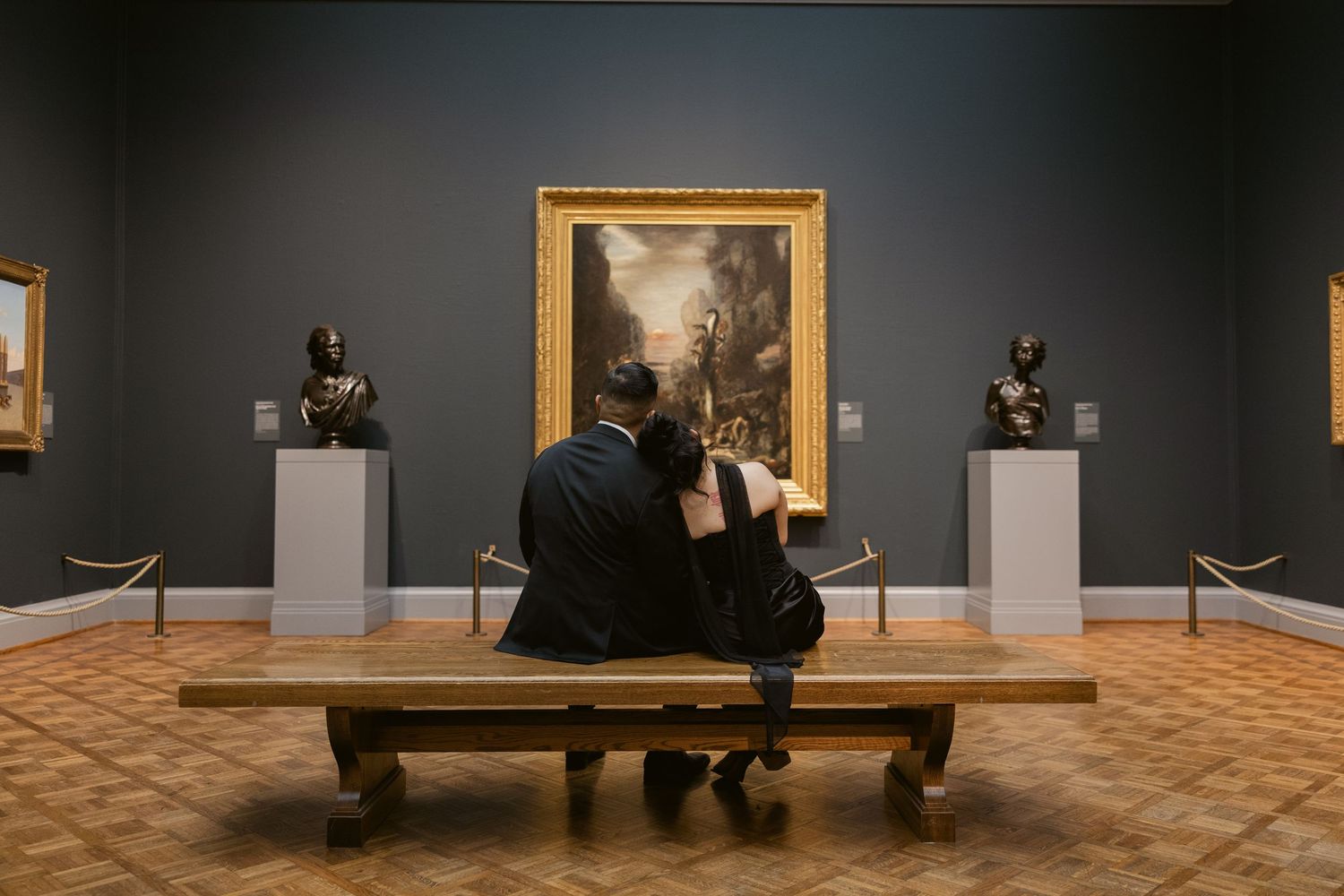 An art gallery interior shows multiple framed paintings on dark walls with wooden flooring.