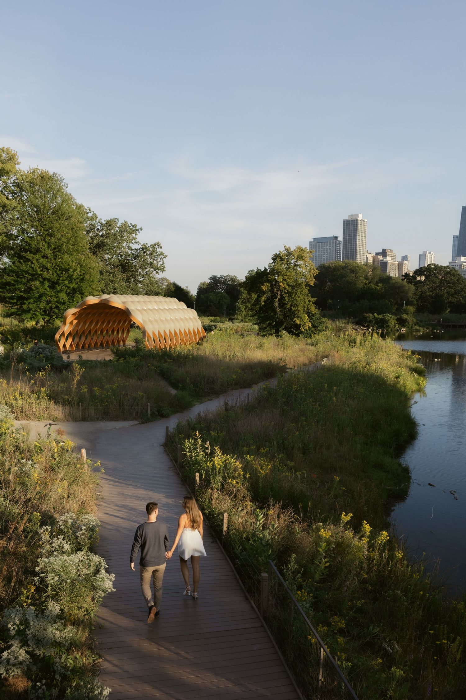 Couple strolling on nature boardwalk with city skyline visible in distance.