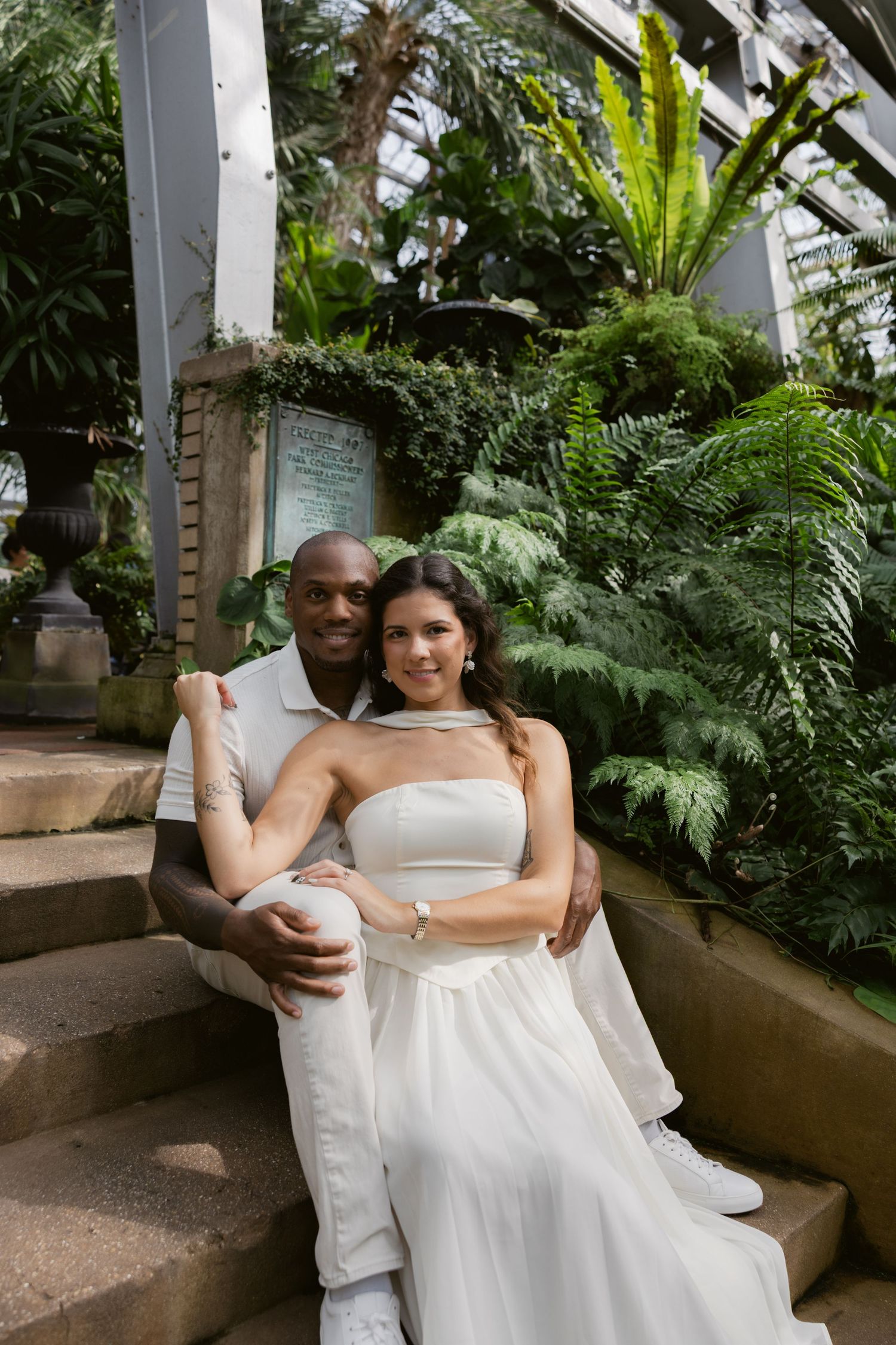 A couple poses together on stone steps surrounded by tropical plants in a greenhouse setting.