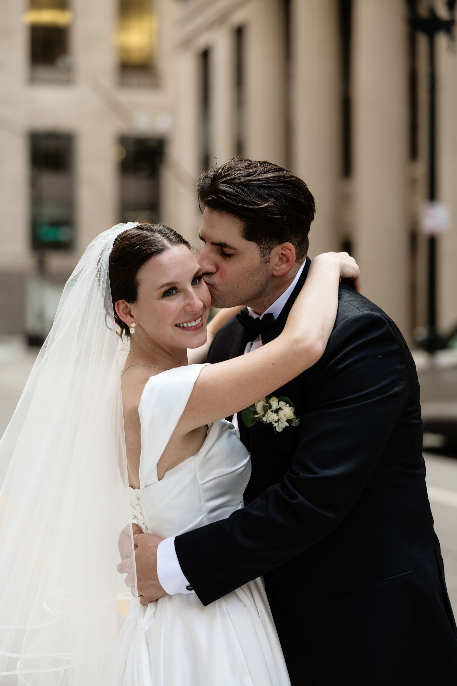 A bride in a white dress and veil embraces her groom in a black tuxedo during an intimate wedding moment.