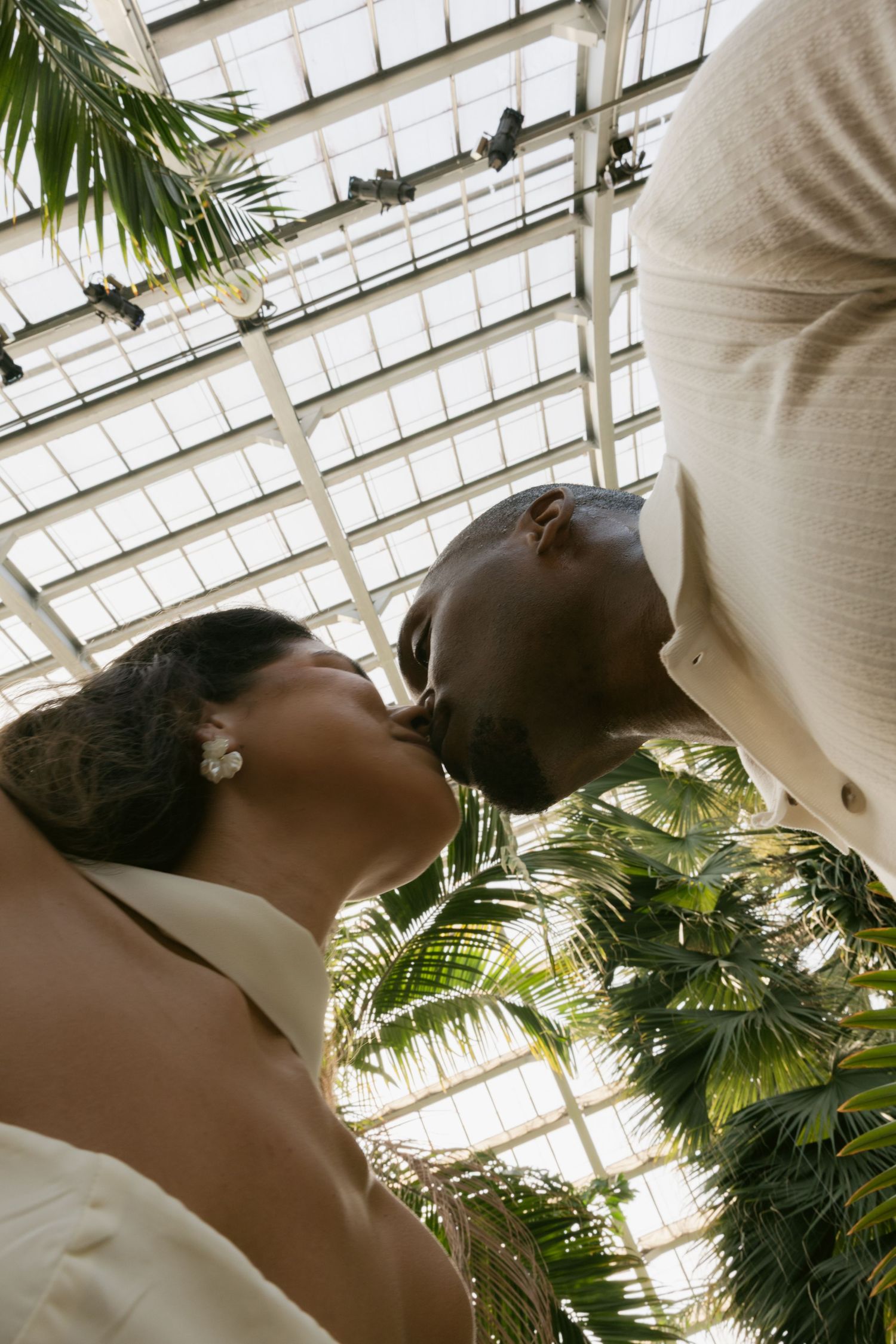 Two people in white attire share an intimate moment beneath a glass greenhouse ceiling surrounded by palm fronds.
