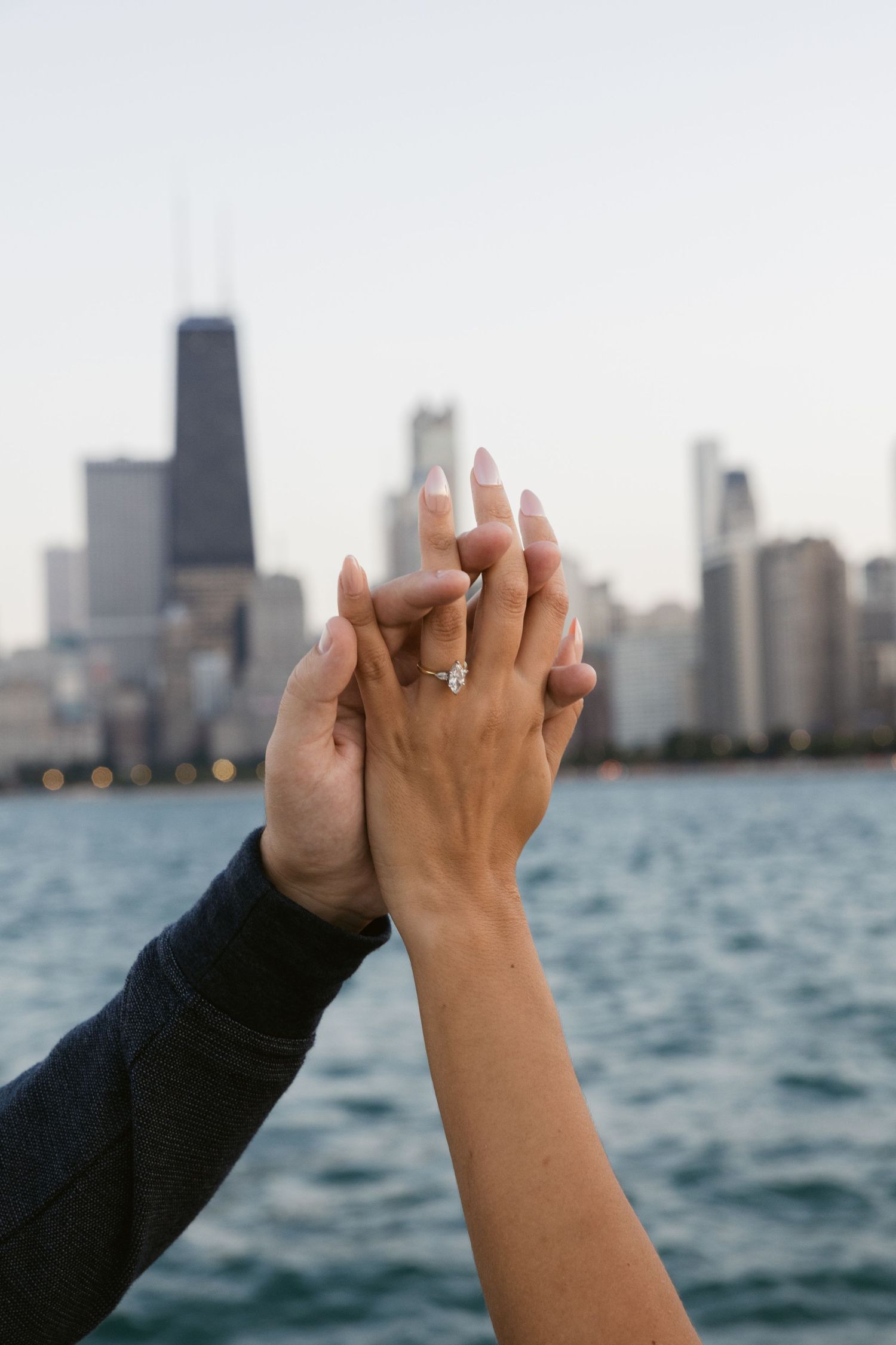 Two hands raised against Chicago skyline showing engagement ring with Lake Michigan in background.
