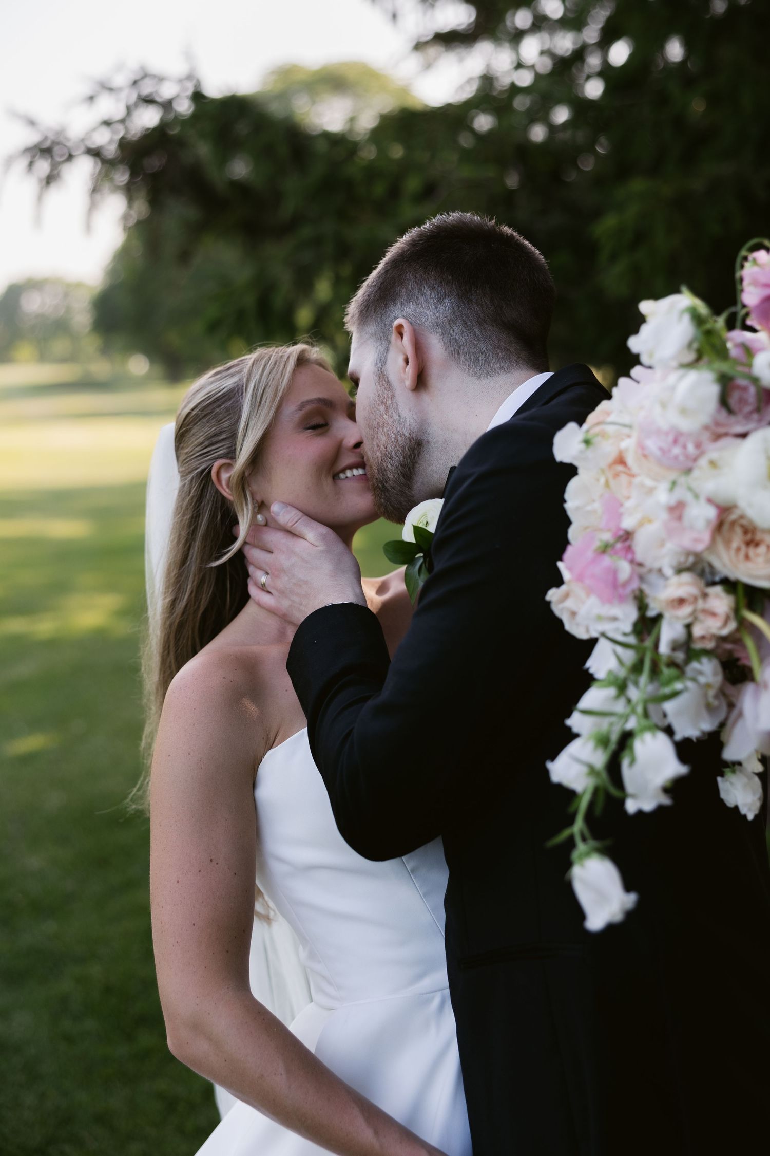 A tender romantic moment between newlyweds next to pink floral wedding decorations outdoors.