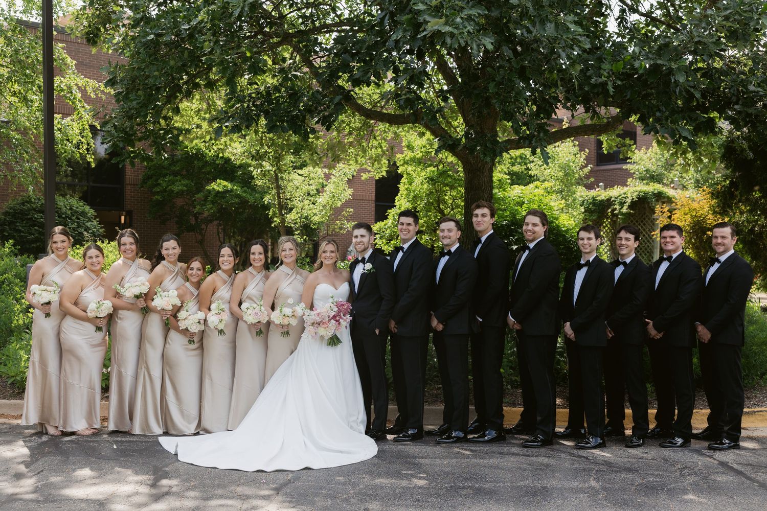 Wedding party members in formal attire pose together outdoors in a garden setting.