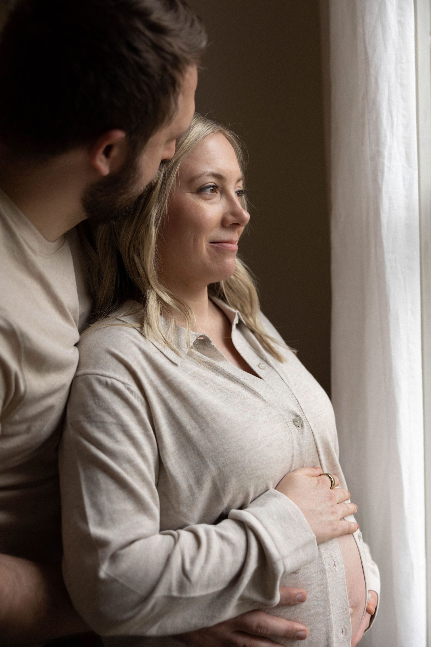 A couple stands by a bright window in tender embrace wearing light colored clothing.