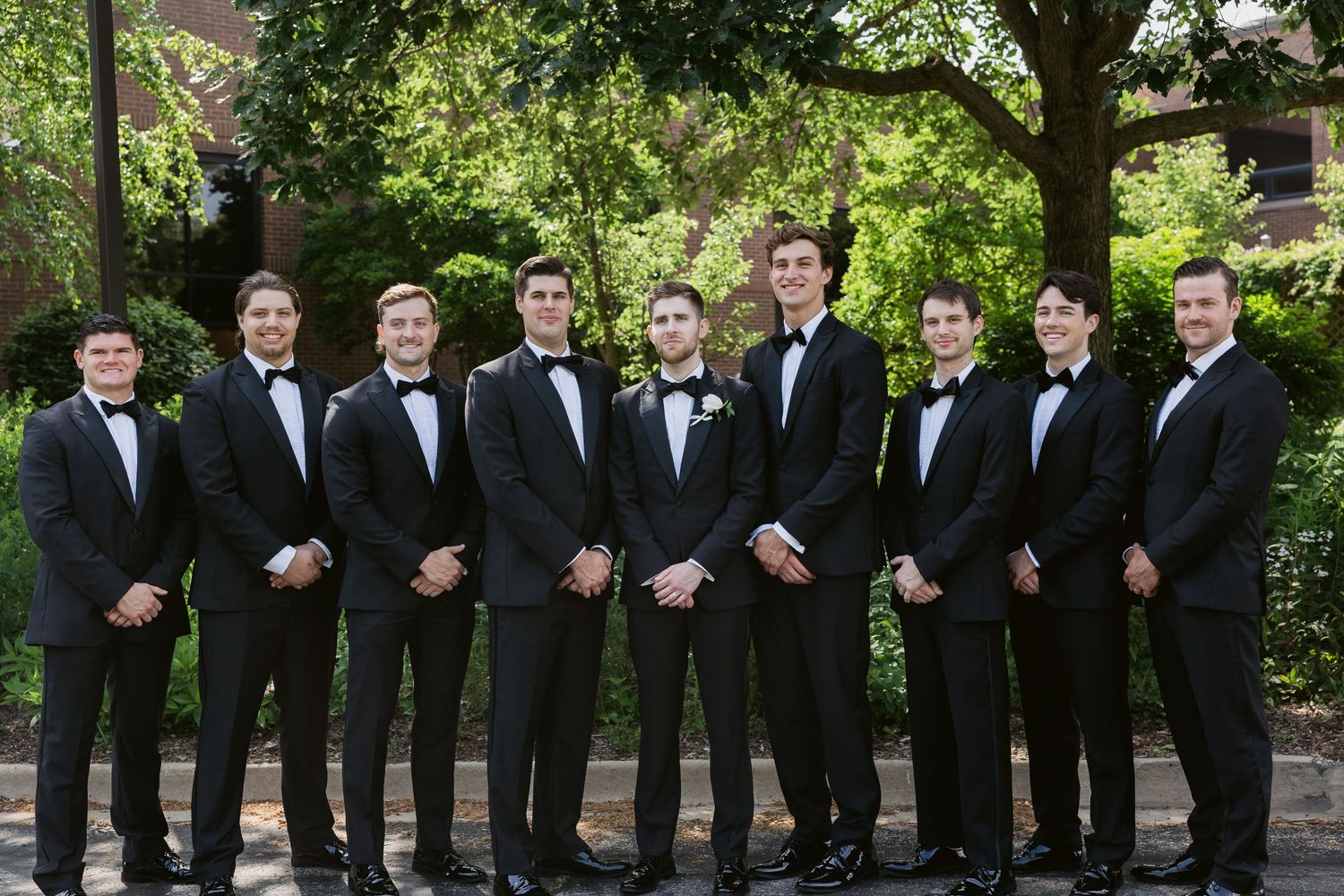 Groomsmen in black tuxedos lined up outdoors for a formal wedding photo.