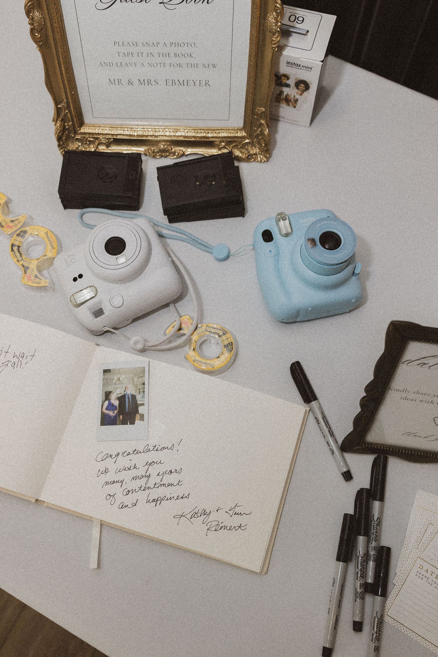 Wedding guestbook with Instax cameras, pens, and decorative gold frame on white surface.