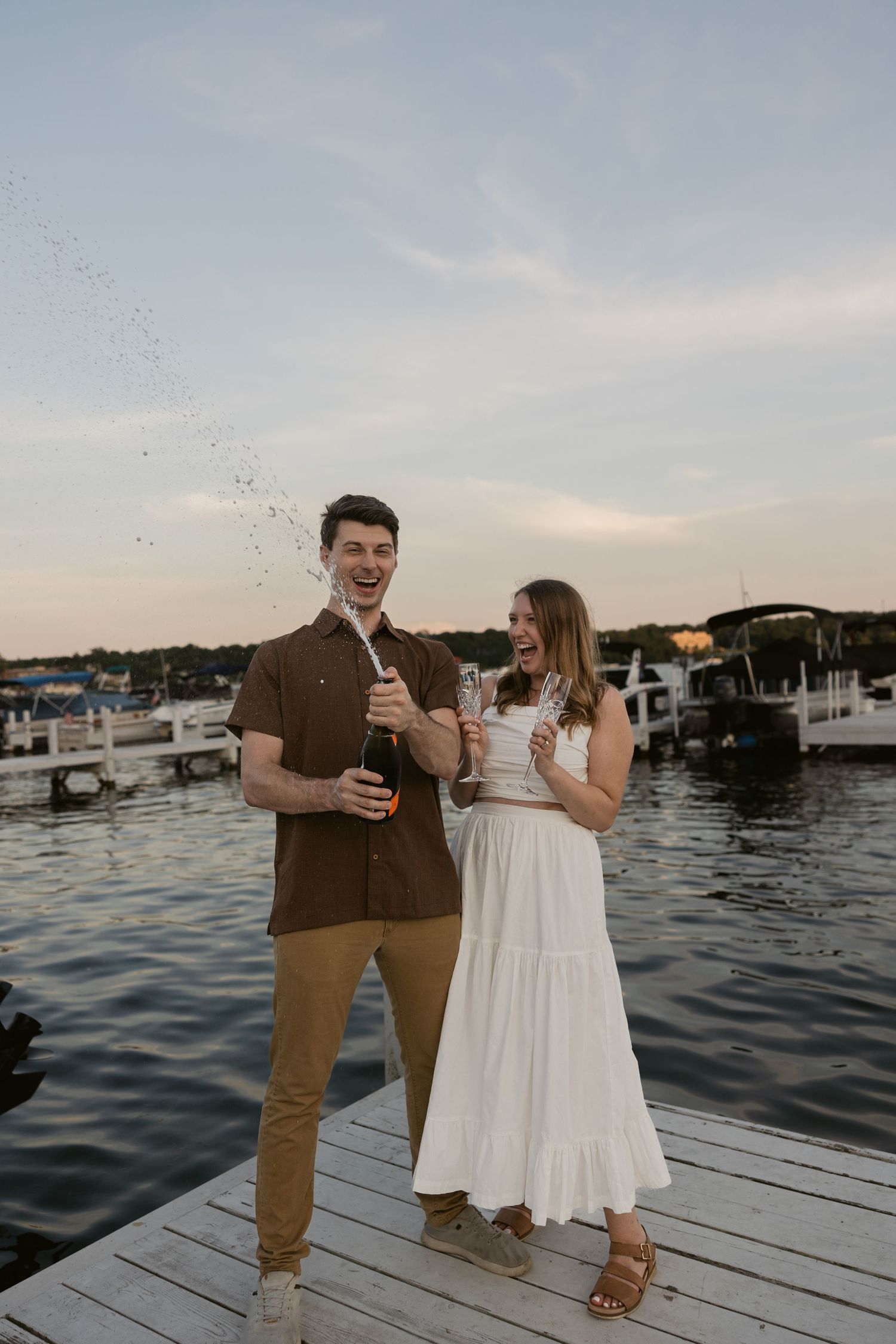 A couple shares drinks and laughs on a waterfront dock during golden hour.