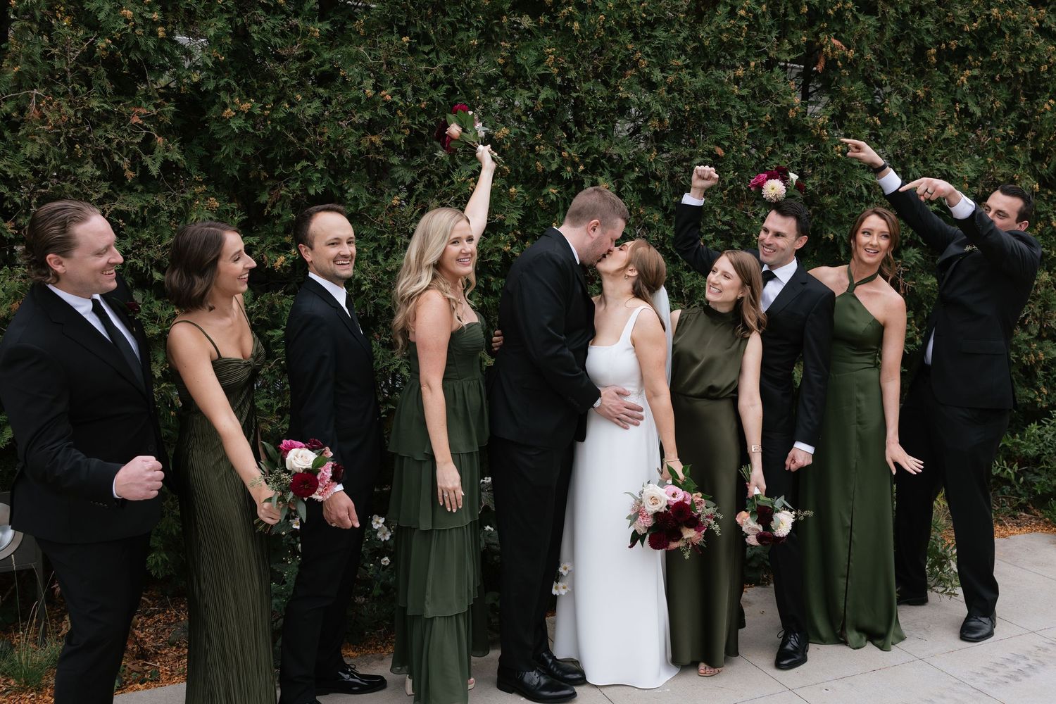 Wedding party in black suits and olive green dresses celebrating with raised arms against hedge wall.