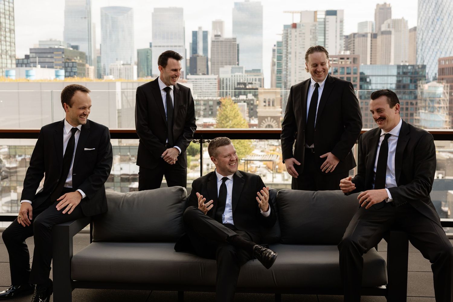 Groomsmen in black suits share laughs and conversation in front of a city skyline backdrop.
