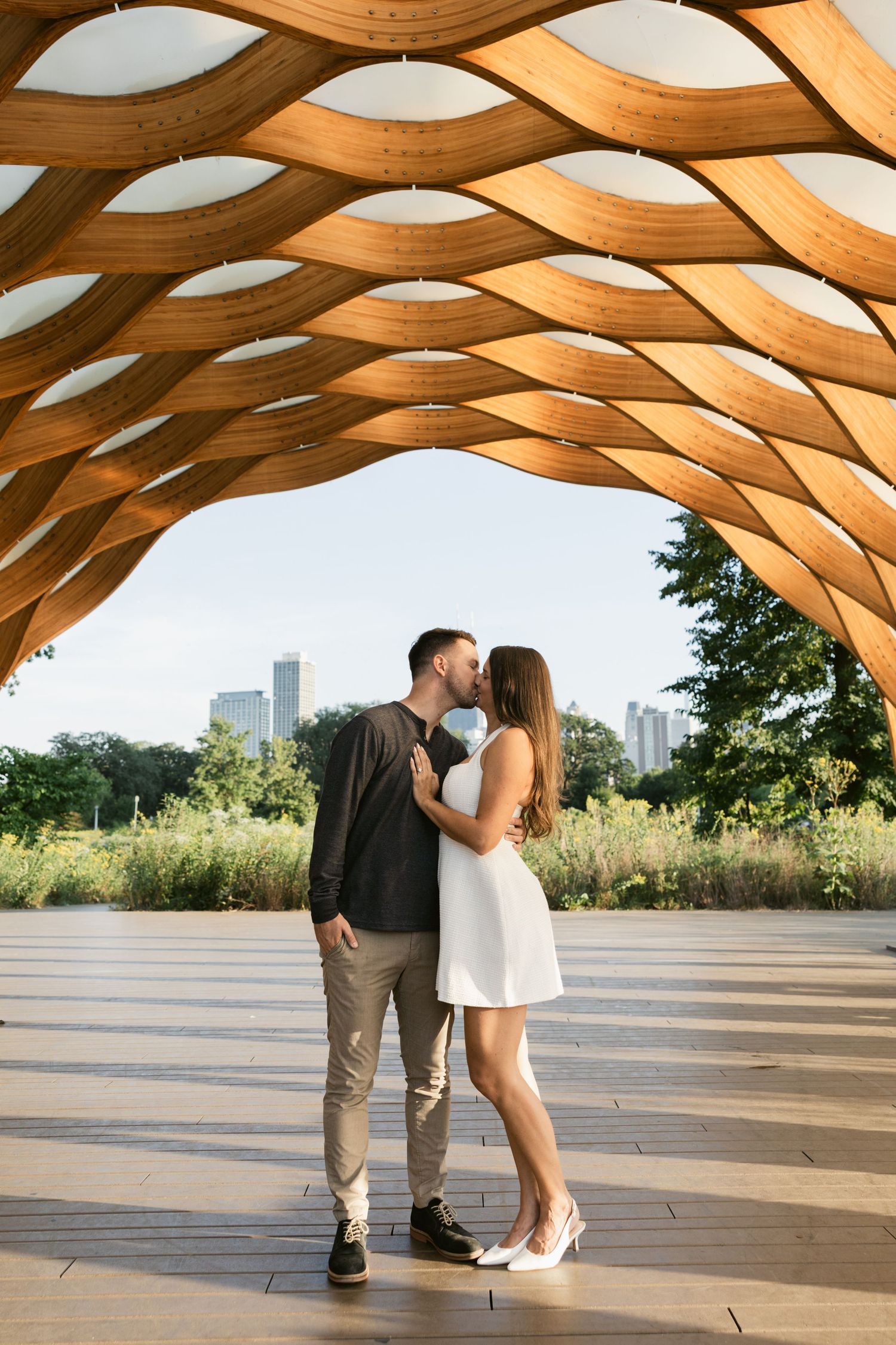 Couple shares romantic moment under wooden lattice pavilion with Chicago skyline visible.