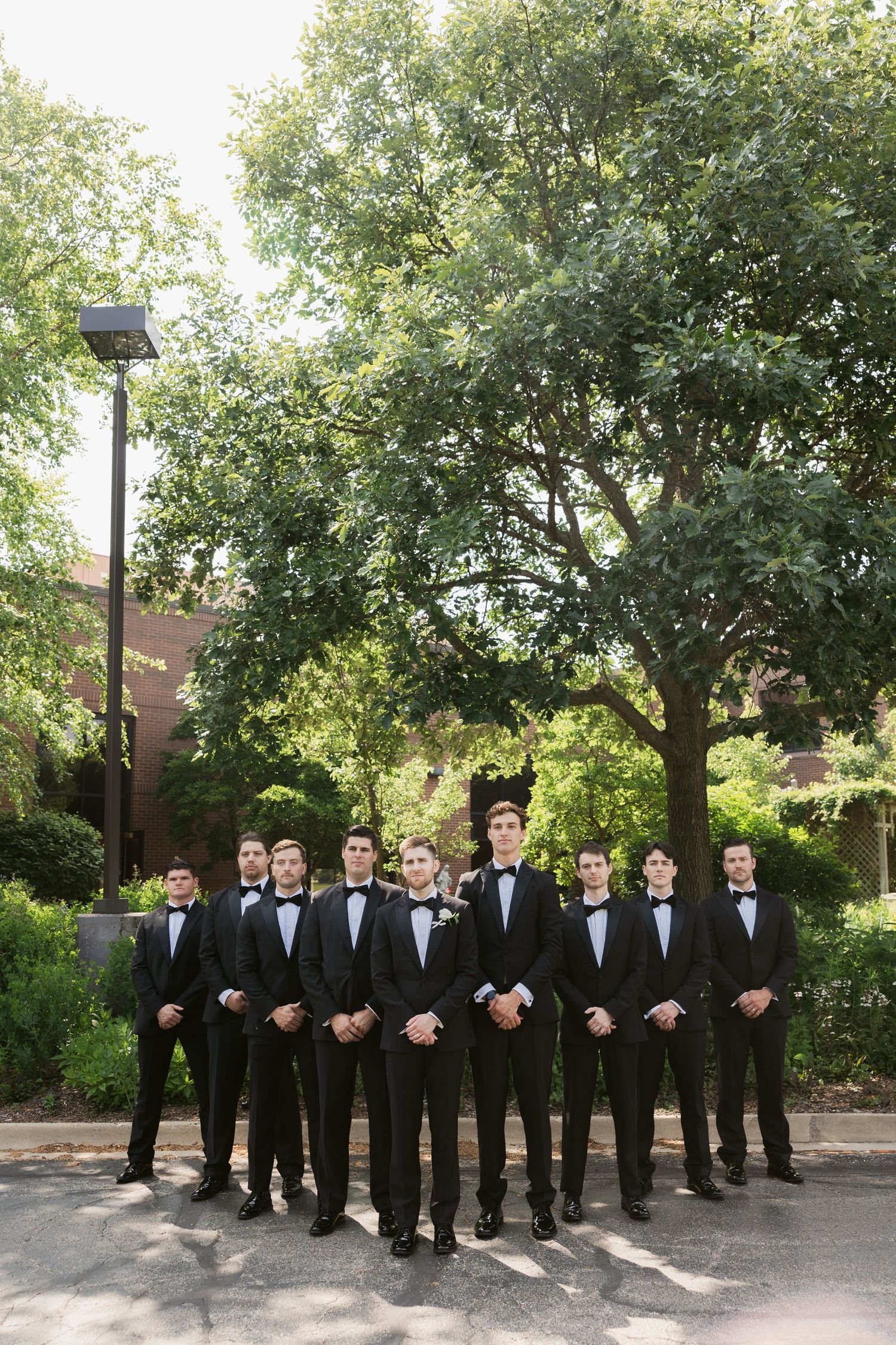 Groomsmen pose in a line wearing black tuxedos in an outdoor garden setting.