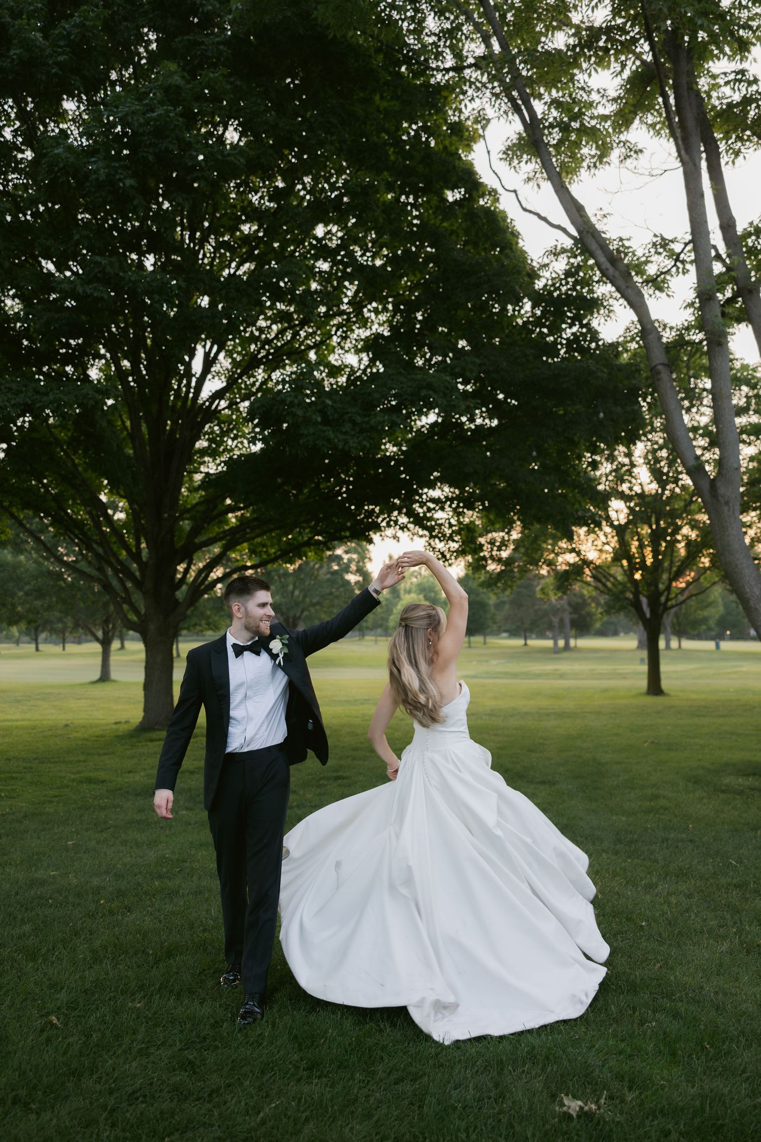 Wedding couple dances playfully on grassy lawn surrounded by trees at golden hour.