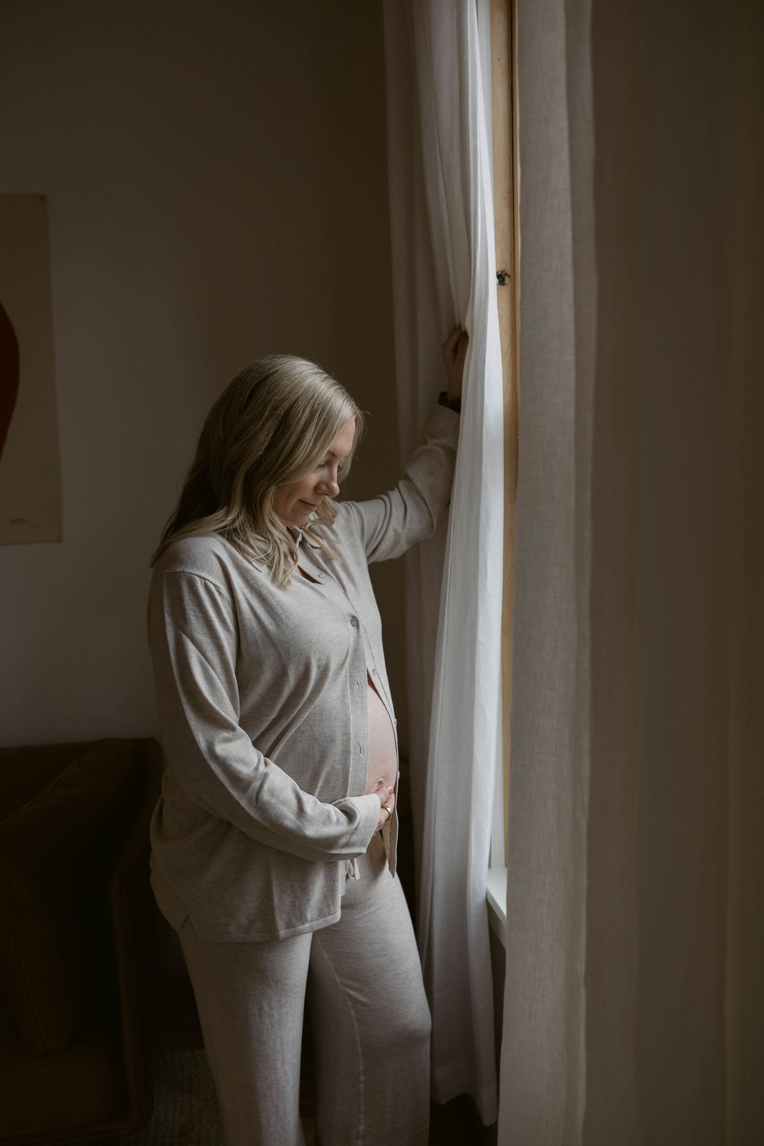 A person in a white sweater stands near a window with soft natural light creating a moody atmospheric scene.
