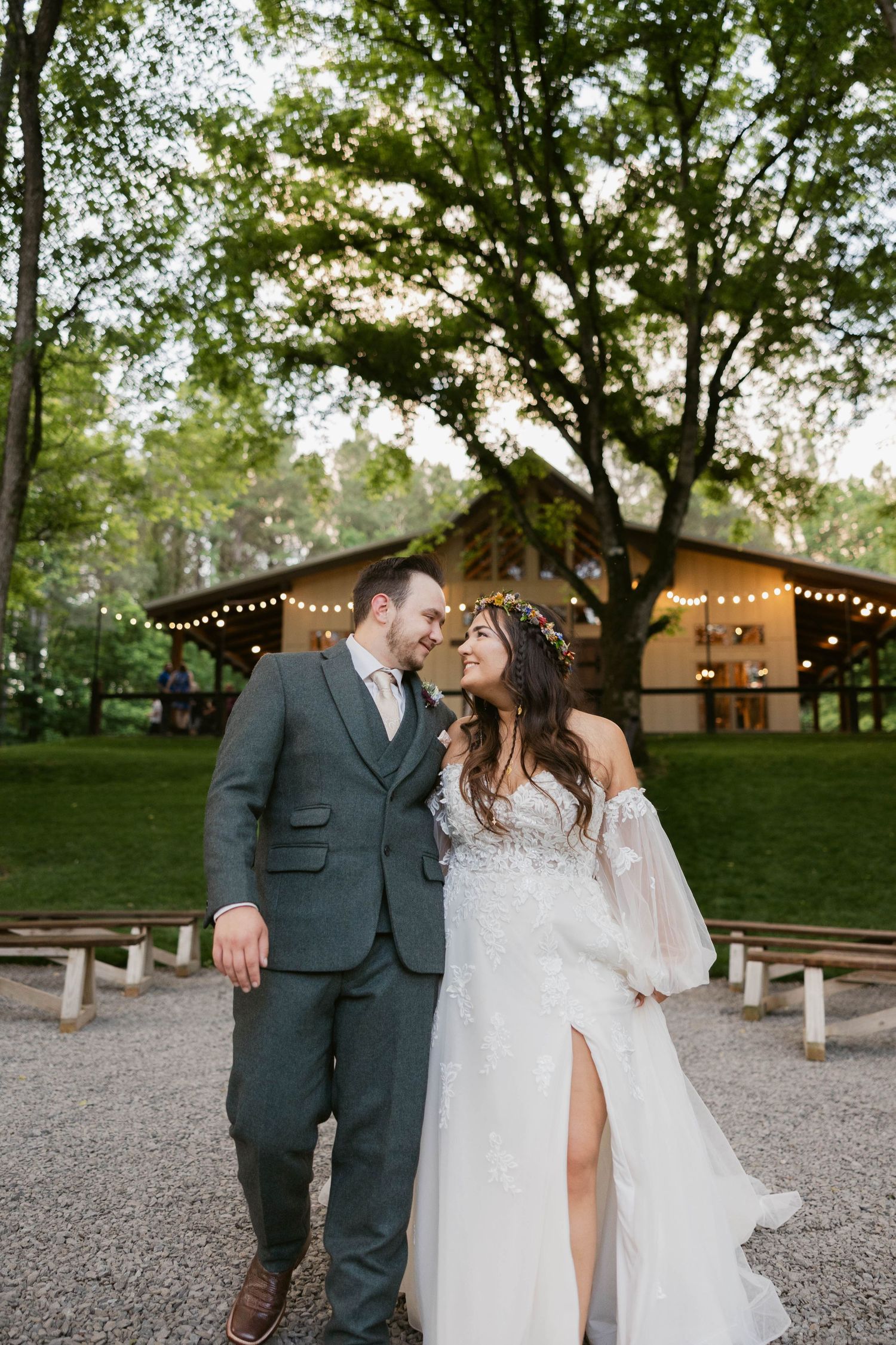 Romantic outdoor wedding photos on gravel path with string lights and rustic venue in background.