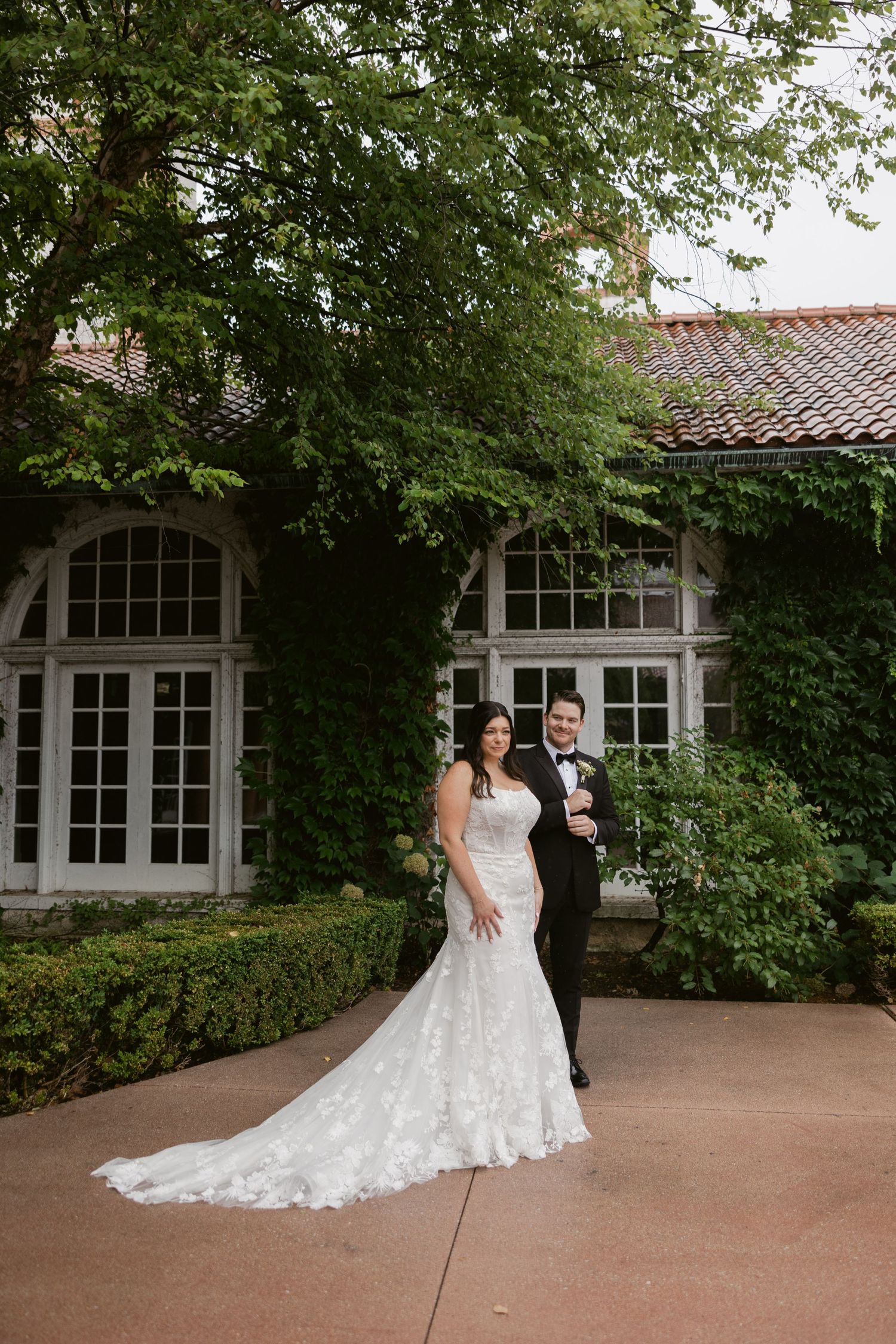 A wedding couple poses in front of a vintage mansion covered in lush green ivy and arched windows.