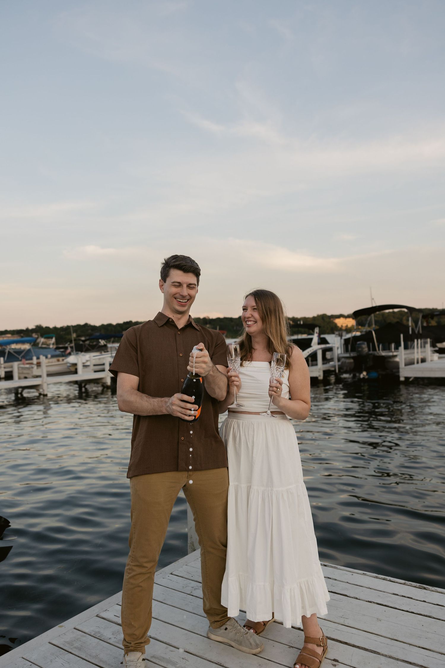 A playful couple in summer outfits dances on a wooden pier with boats in the background.