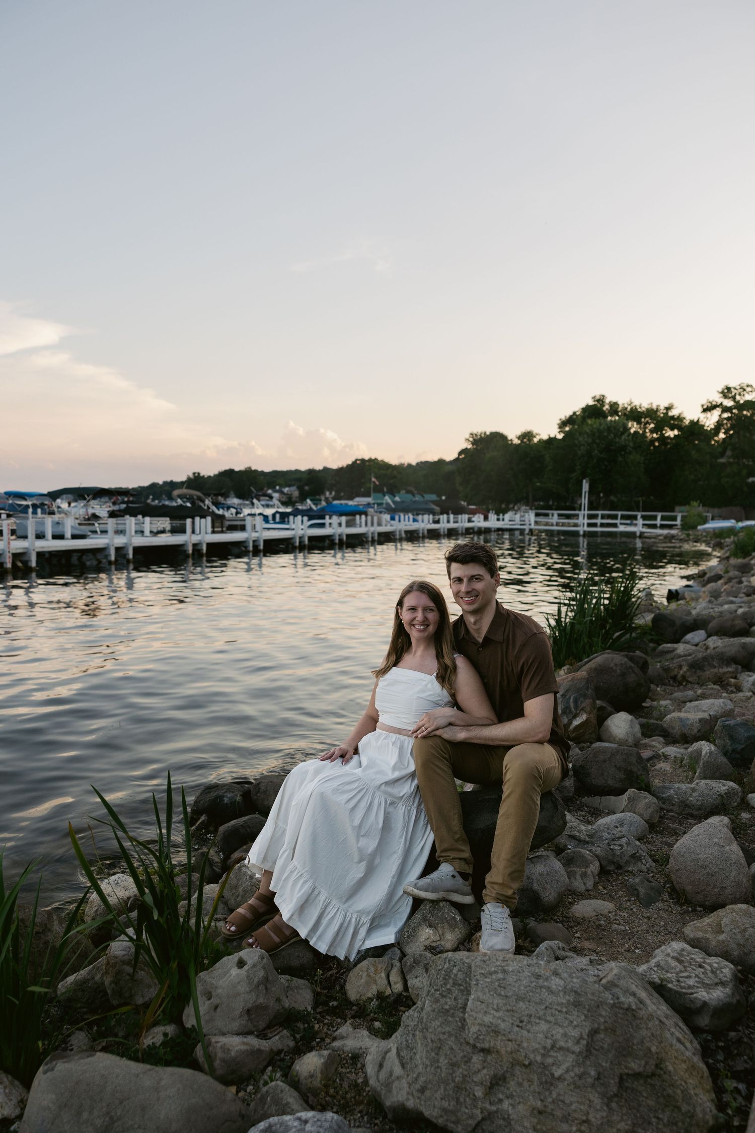 A couple sits together on rocky shoreline with marina and boats visible in the sunset background.