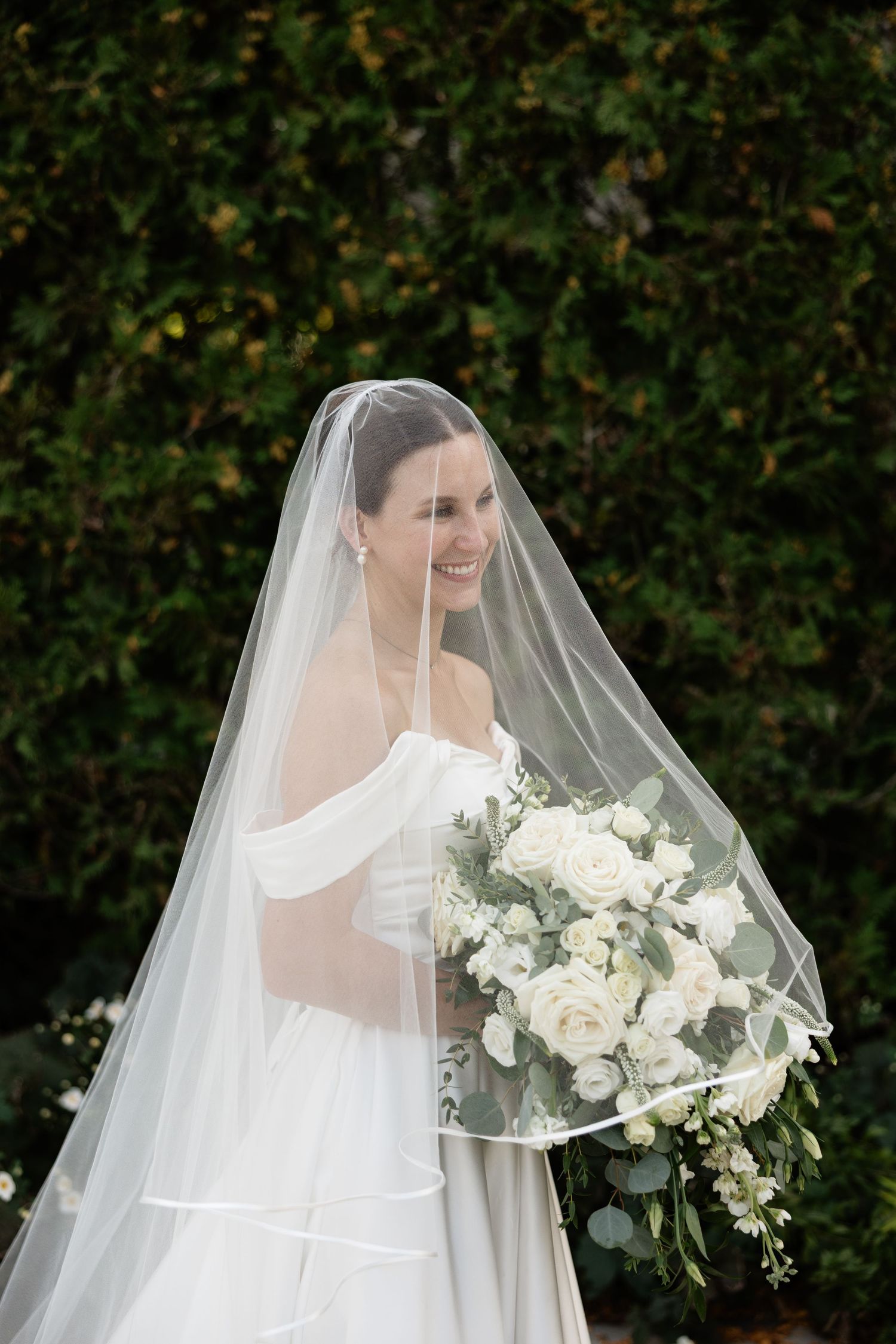 A bride in a white off-shoulder wedding gown holds an elegant bouquet of white roses and greenery against a garden backdrop.