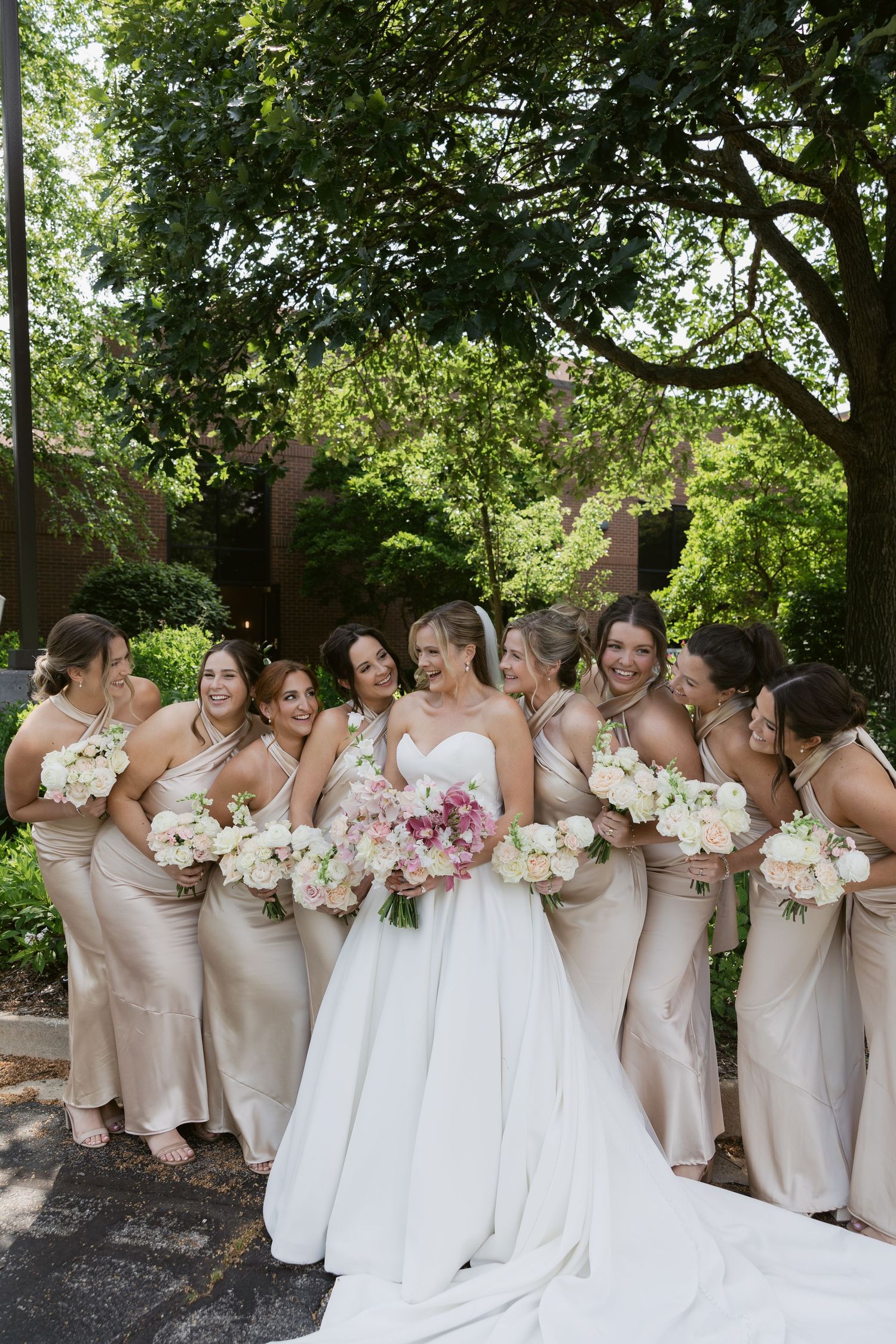 A bridal party in blush dresses poses outdoors with bouquets of pink and white flowers.