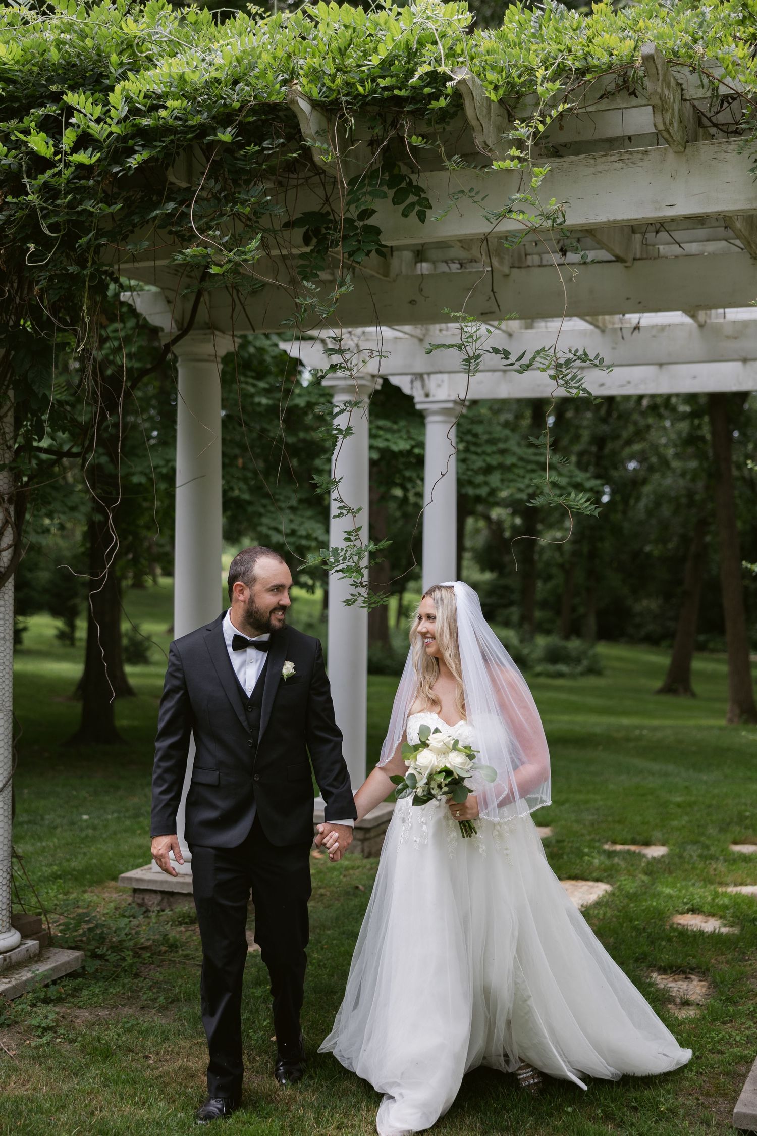 A couple celebrates their wedding day beneath a white pergola covered in lush greenery in a garden setting.