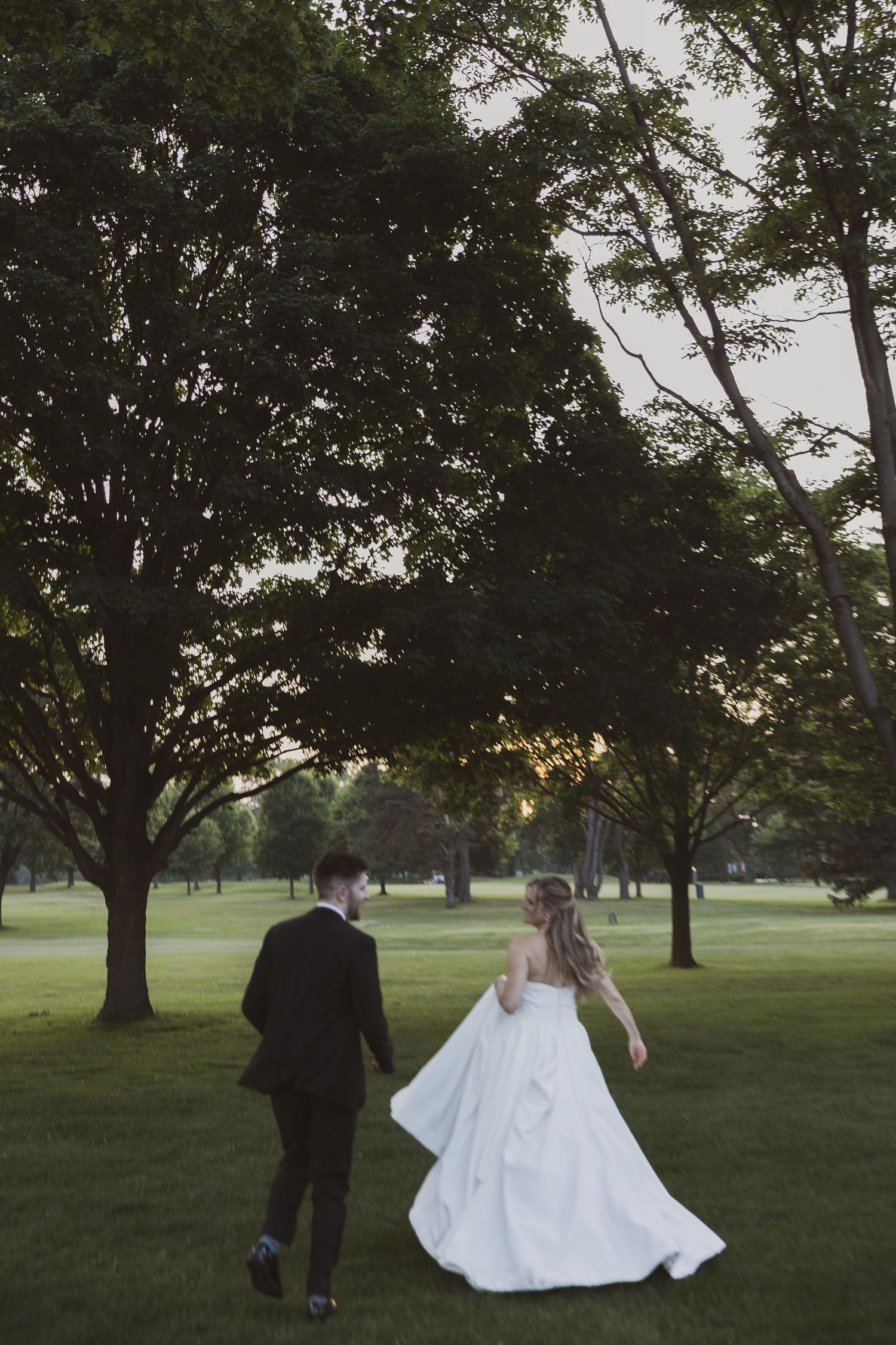 Newlyweds walking together across a scenic green lawn with large trees at sunset.