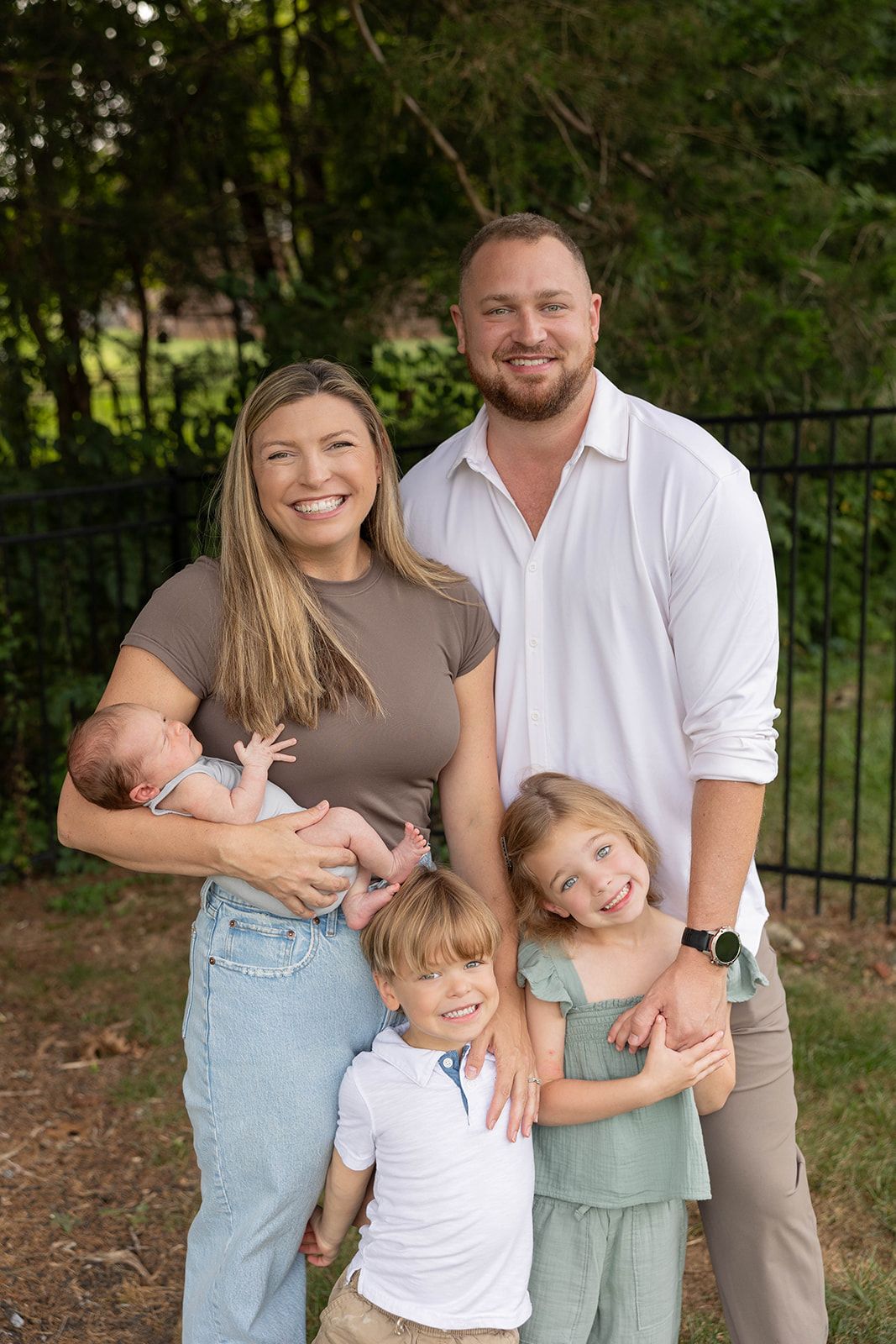 Happy family of five posing together for outdoor portrait in front of trees and fence.