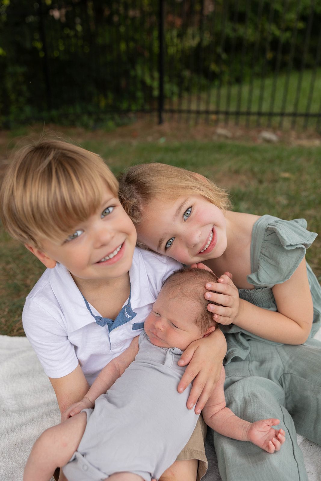 Two children on a blanket embracing a baby outdoors by a garden fence.