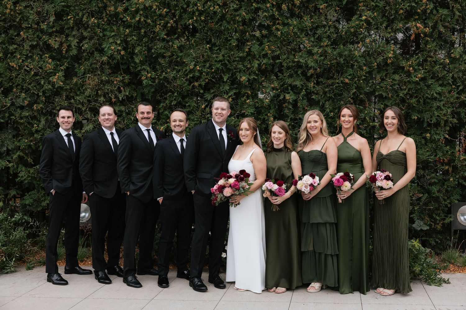 Wedding party poses in formal attire with bridesmaids in olive green dresses and groomsmen in black suits.