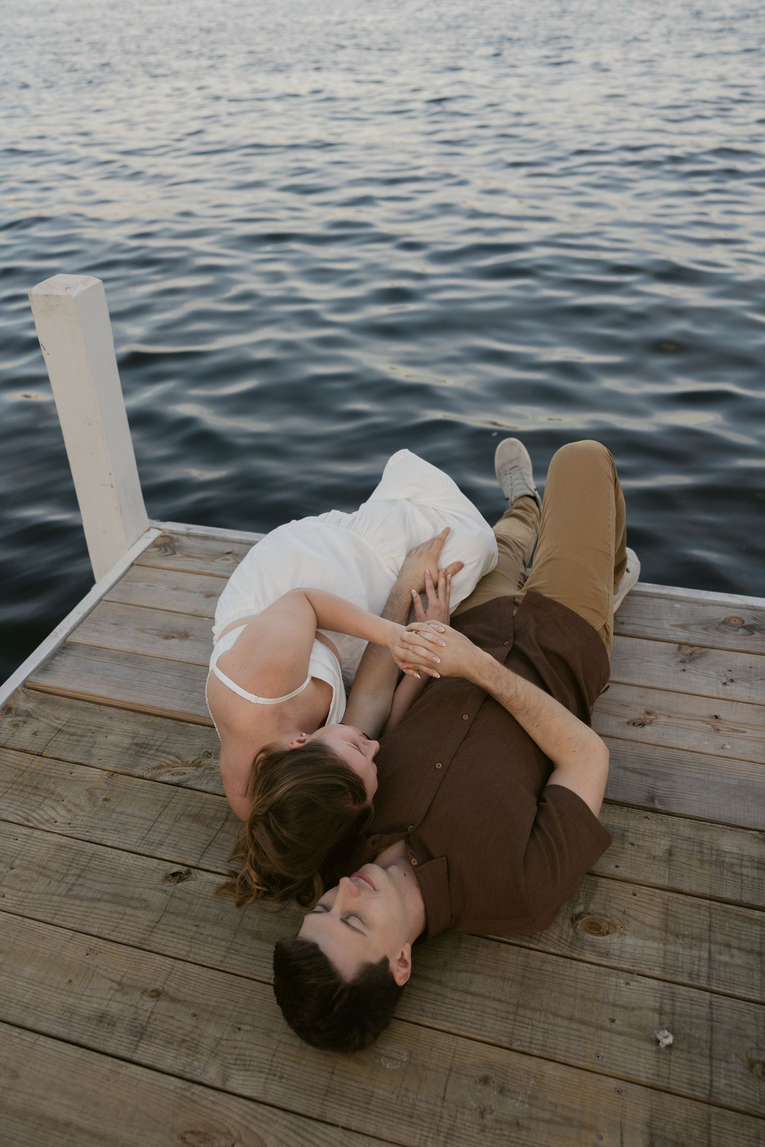 A couple lying together on a wooden dock over calm blue water during a lakeside photoshoot at sunset.