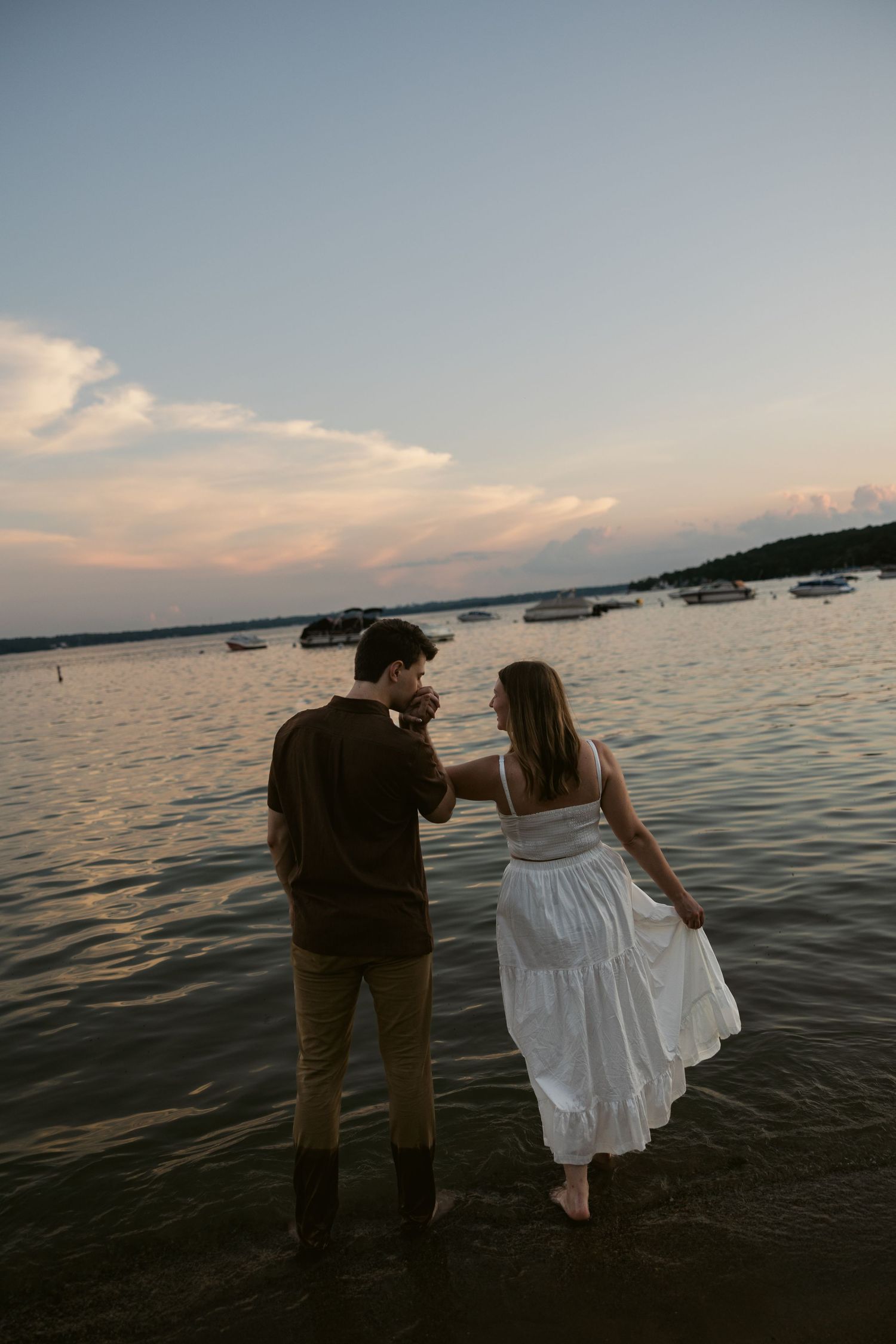 A romantic silhouette of a couple stands at the edge of a calm lake at sunset with boats in the background.