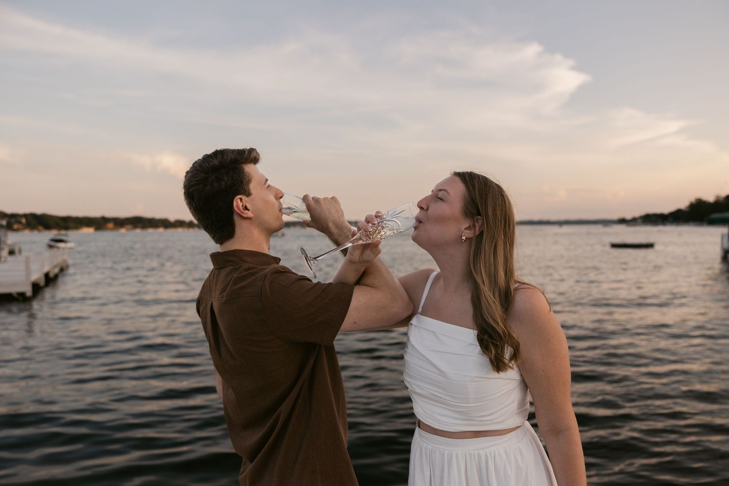 Playful couple drinking champagne together on dock at sunset.