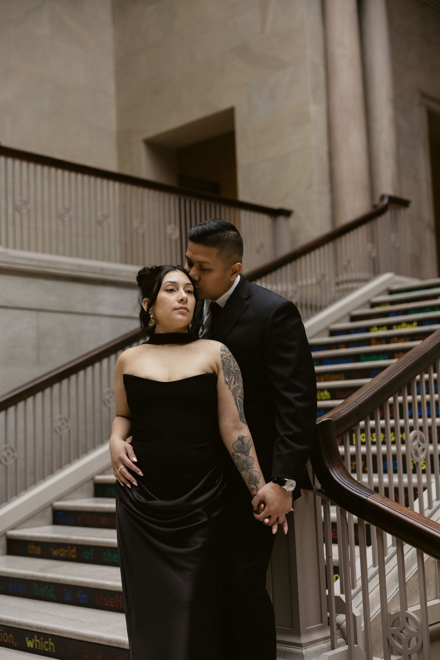 A romantic couple in black formal attire embraces in an elegant architectural setting.