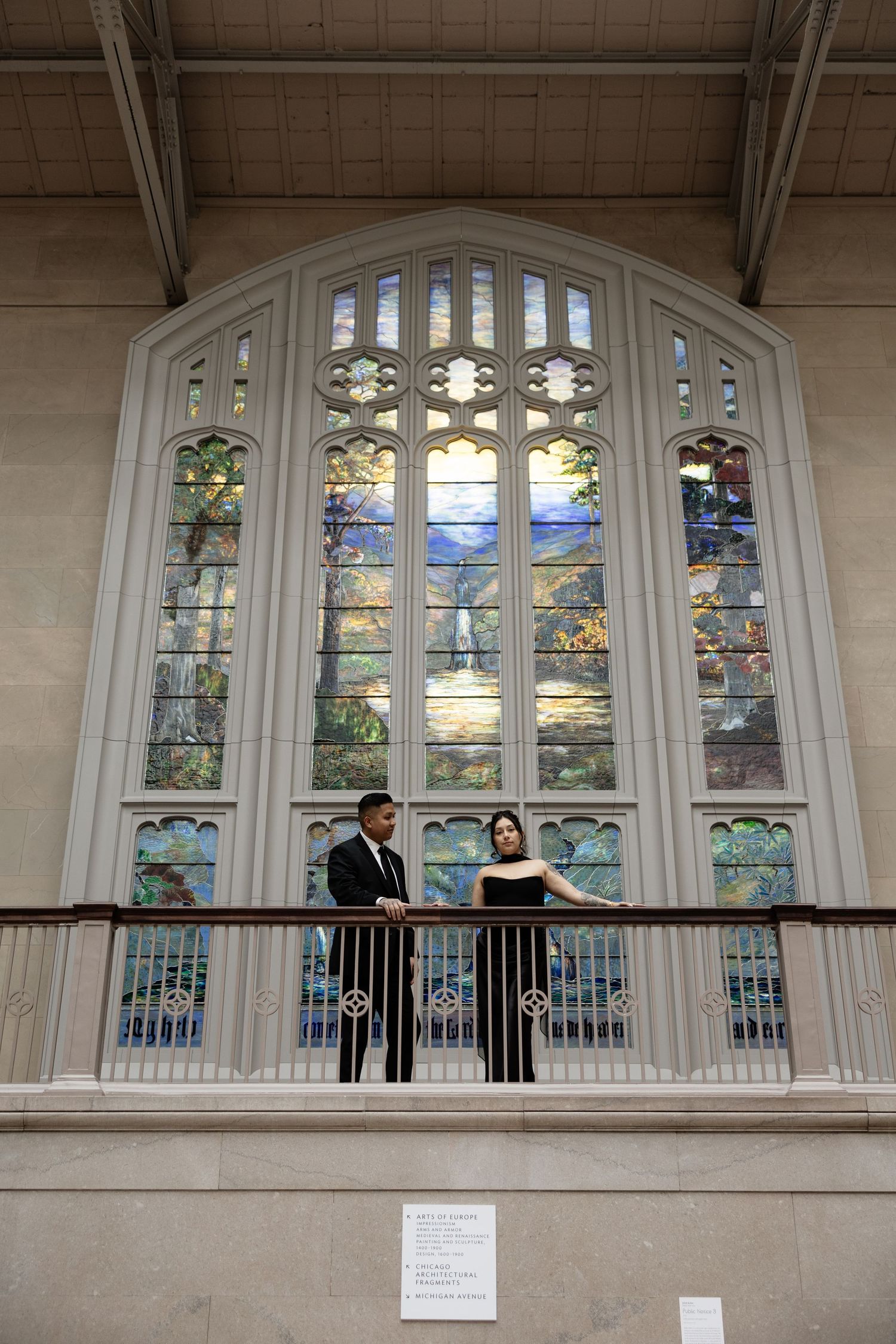 A couple stands on a balcony with beautiful stained glass windows in an elegant architectural setting.
