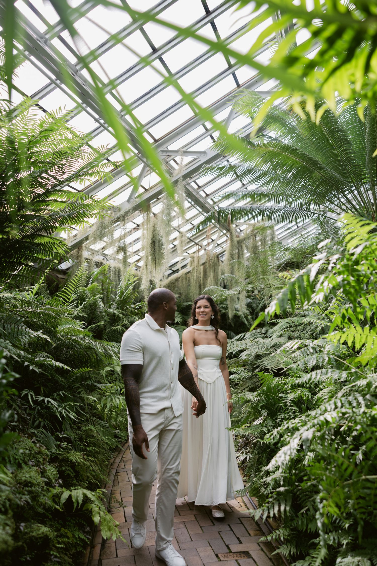 A couple walks together on a brick path through a verdant greenhouse filled with ferns and tropical plants.