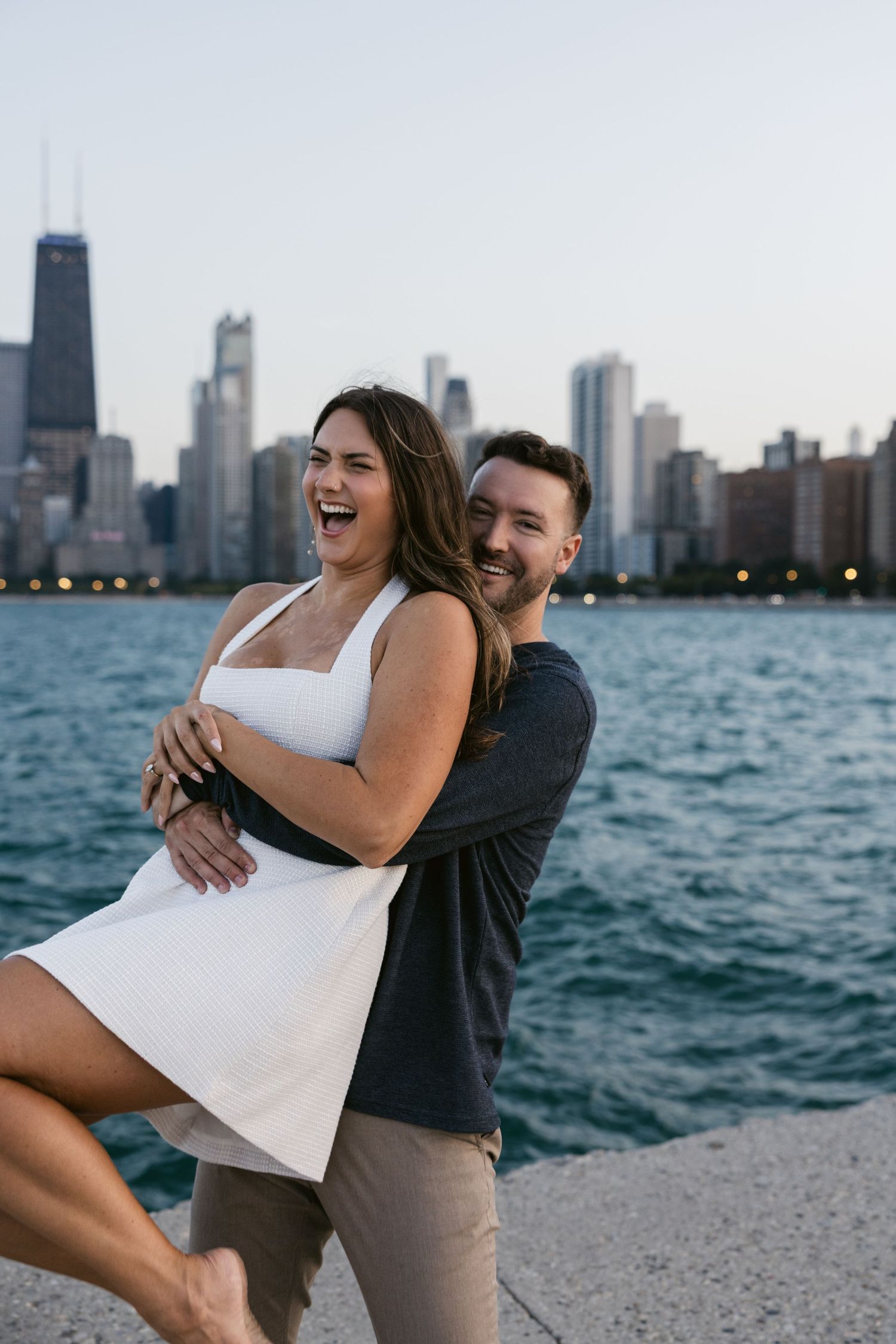 Joyful couple laughing and embracing by Lake Michigan with Chicago's skyline at dusk.