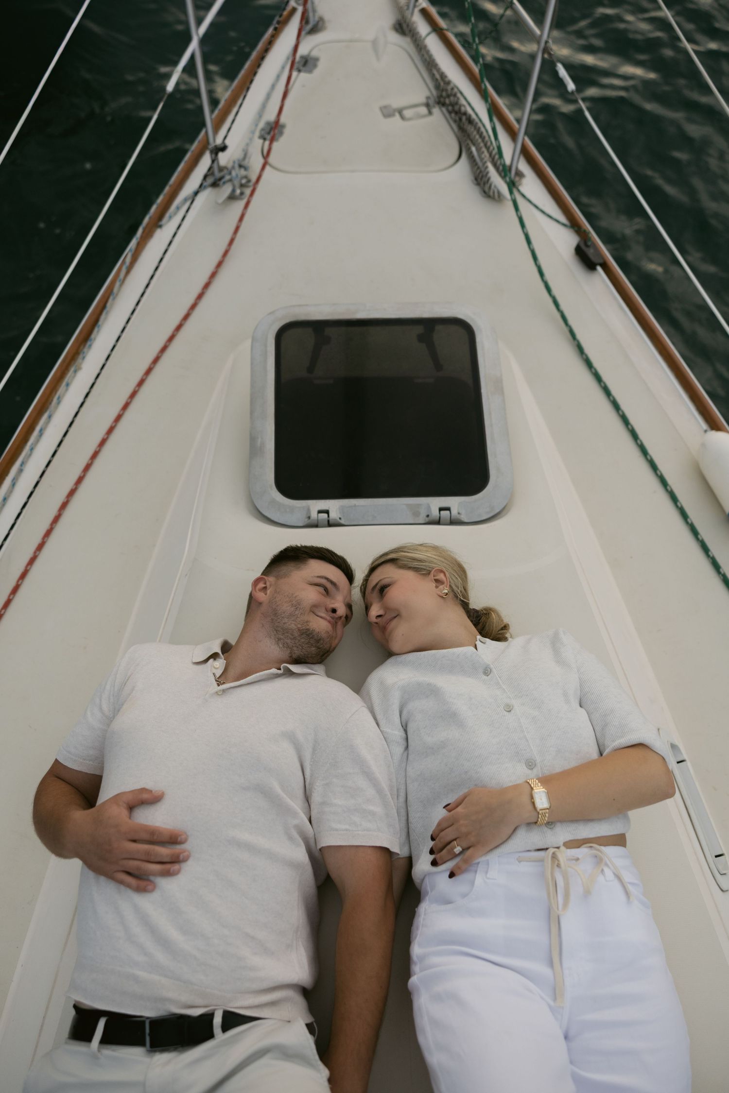 Couple in white outfits lying together on the deck of a sailboat with city skyline in background.