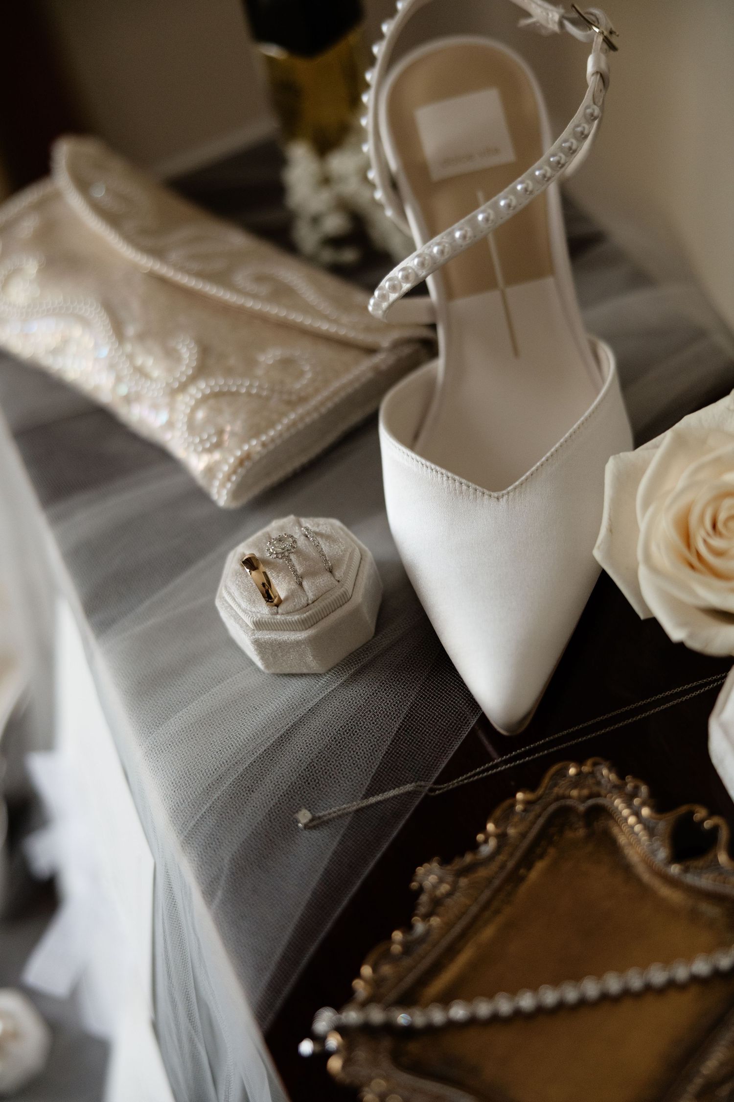 Elegant white bridal shoes with pearl details arranged with delicate roses and jewelry accessories on a soft gray surface.