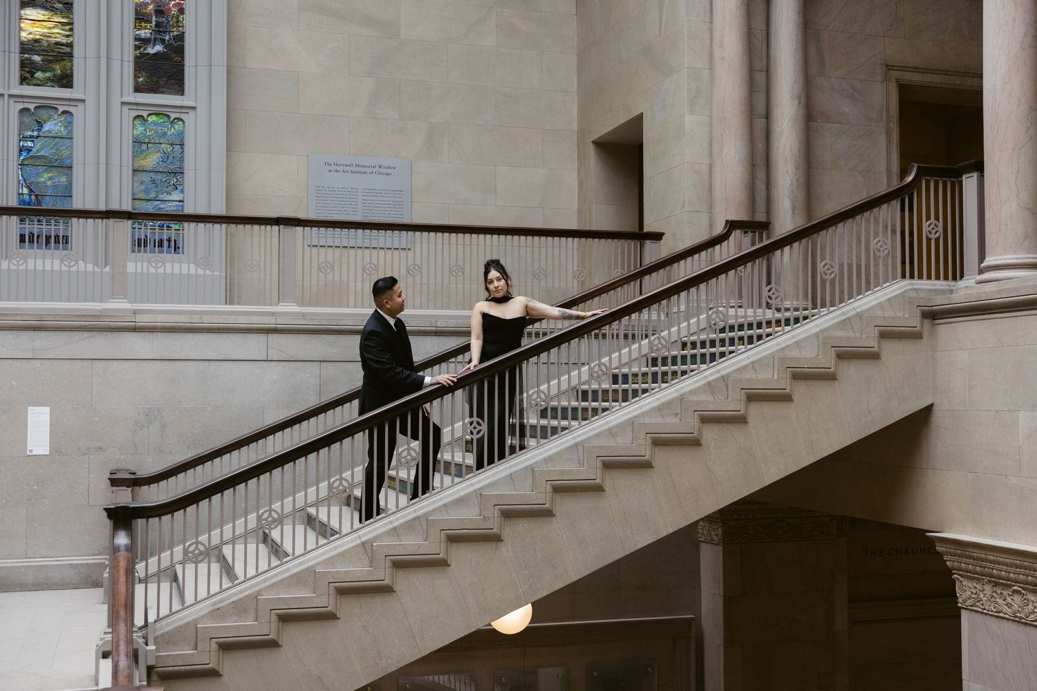 Two figures stand on an elegant stone staircase with ornate railings inside a grand historic building.