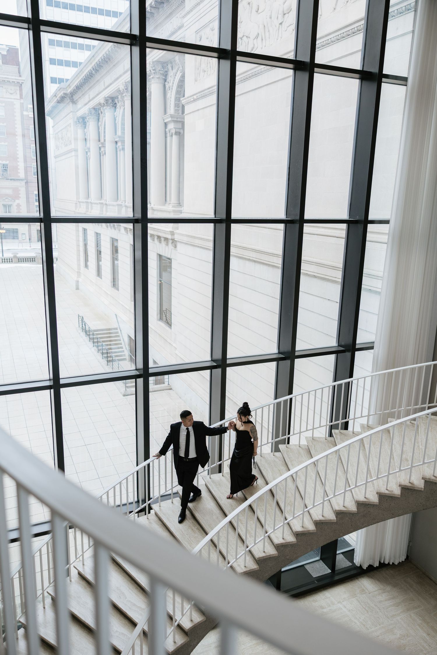 A couple walks along a modern curved staircase with large windows and white railings.
