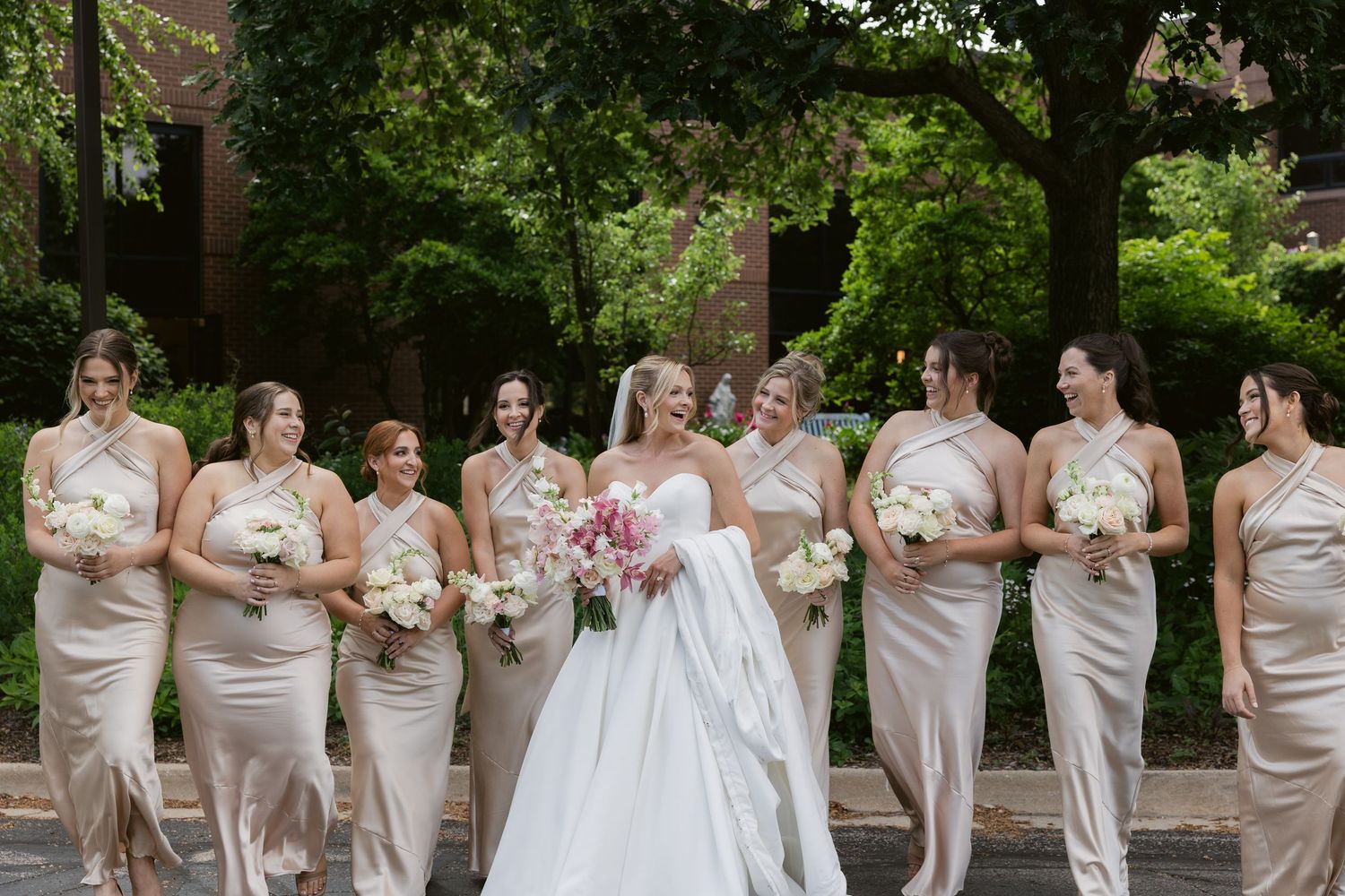 A wedding party in champagne colored bridesmaid dresses stands together for a formal outdoor portrait.