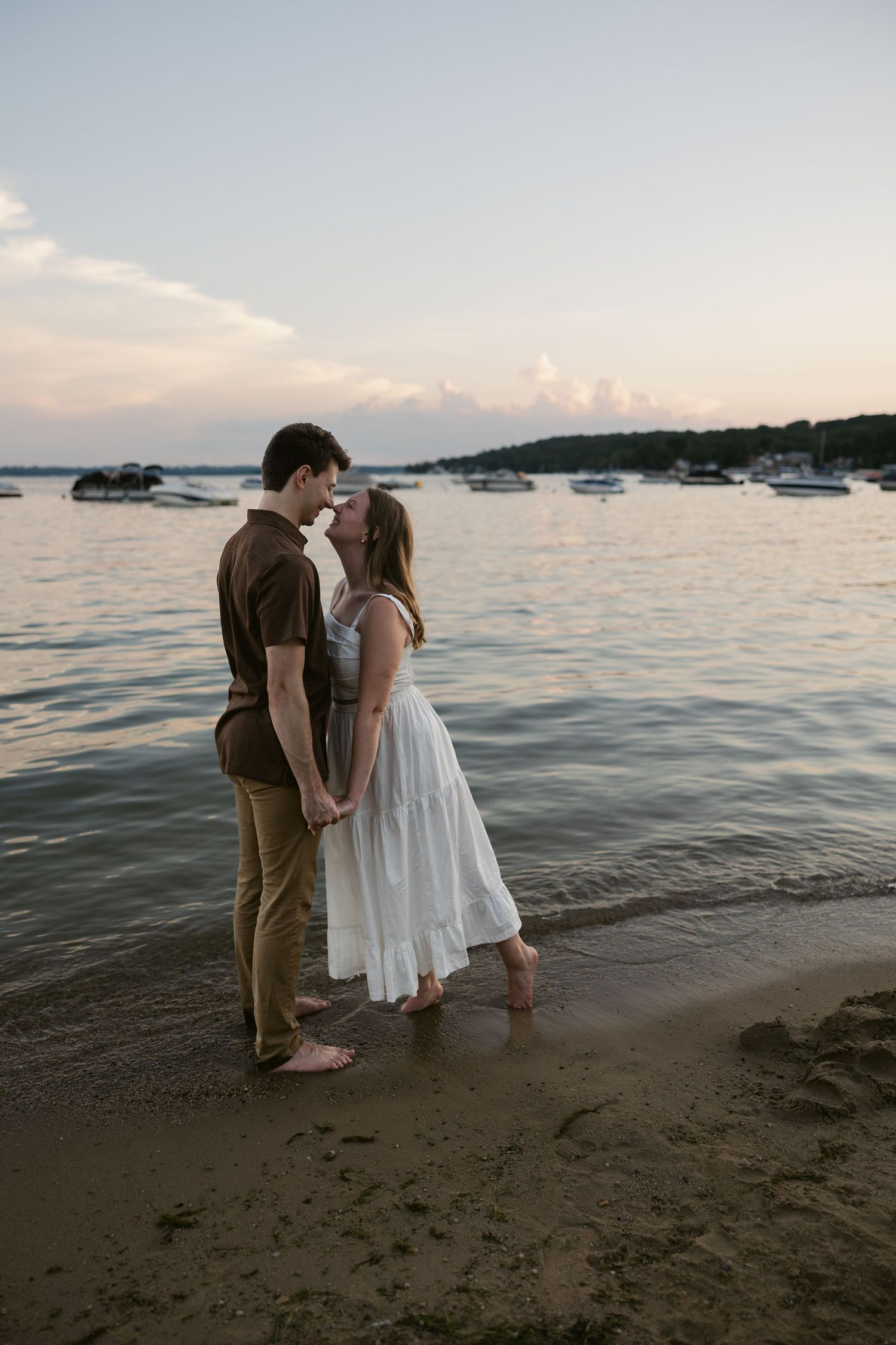 Two people in white and brown outfits embracing on a beach with calm waters in the background.