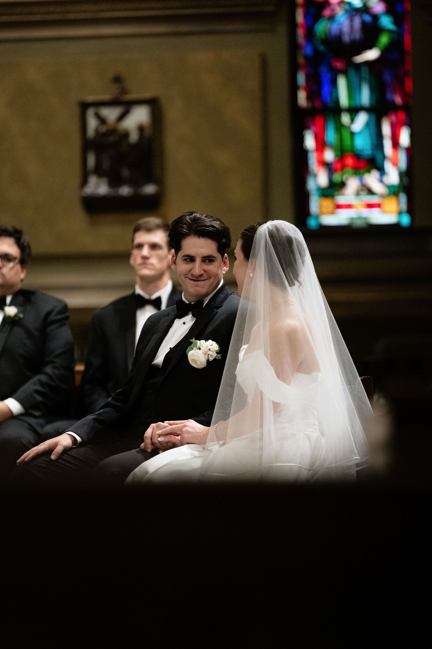 A couple sits together during a church wedding ceremony with stained glass windows visible in the background.